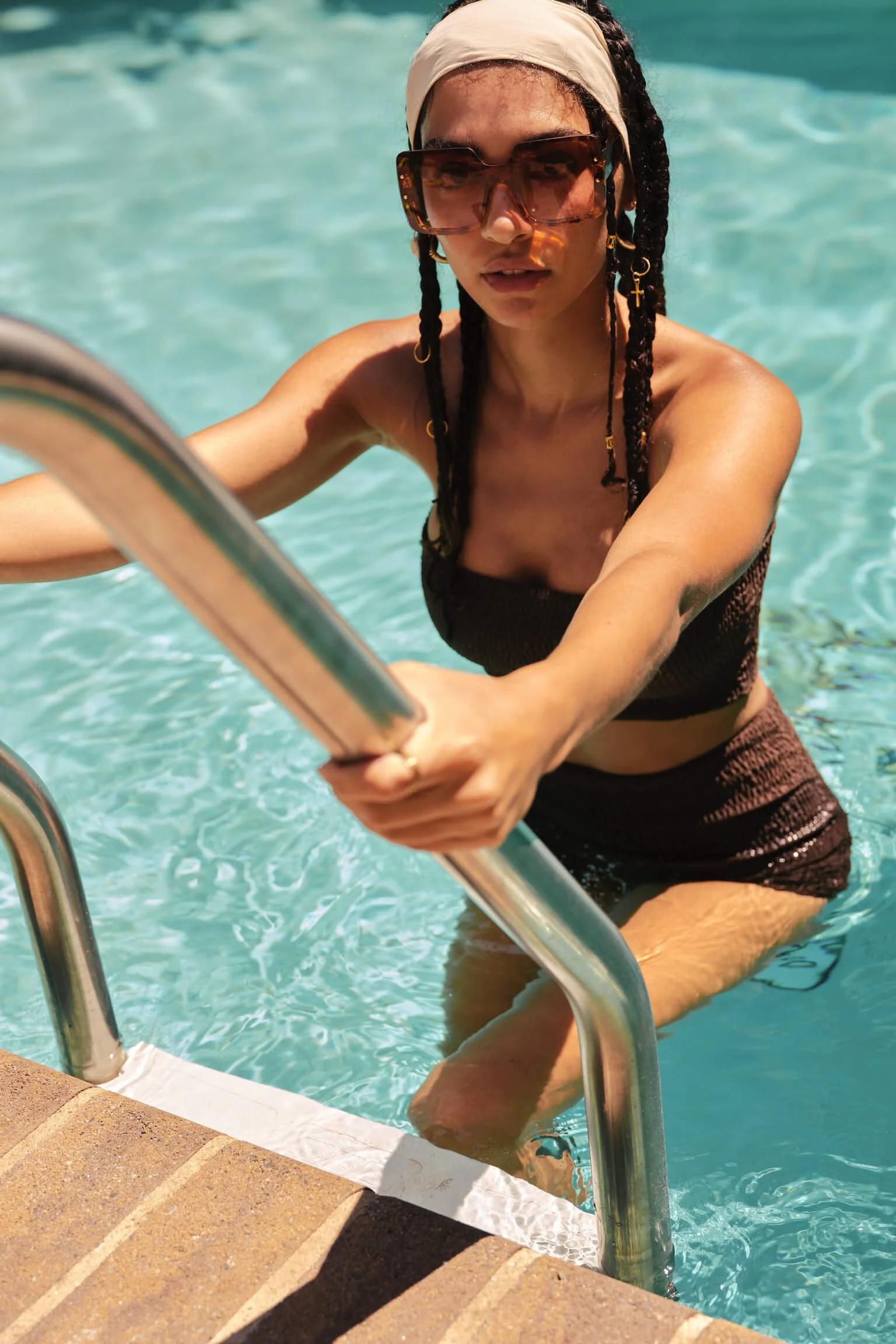 A woman with dark braided hair, wearing large sunglasses, a white headband, a black swimsuit, and dark shorts, climbing out of a swimming pool on a metal ladder.
