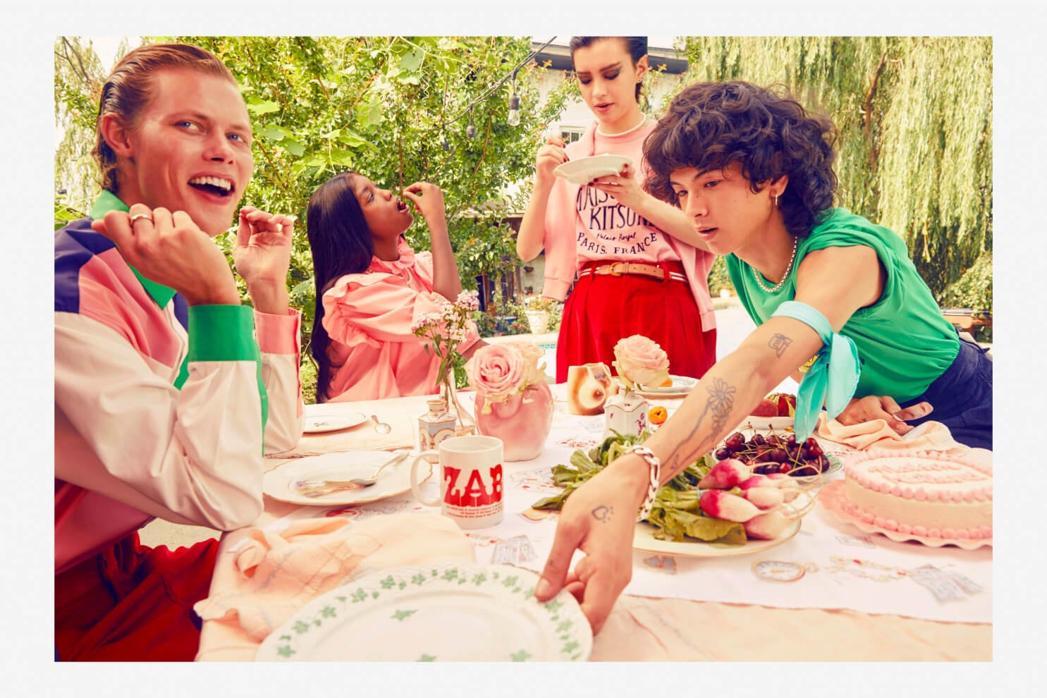 Group of five friends sitting outdoors at a decorated table with flowers and food, enjoying a casual gathering on a sunny day.