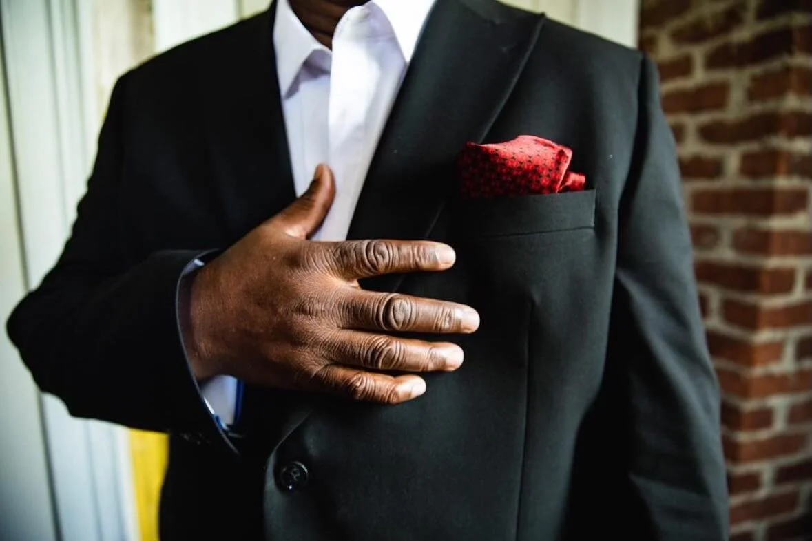 Close-up of a man wearing a black tuxedo with a white shirt and a red pocket square, placing his right hand over his heart with a brick wall background.