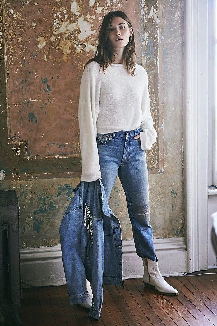 Young woman standing indoors, wearing a white sweater, blue jeans, and white boots, holding a denim jacket, with a rustic textured wall and a window in the background.
