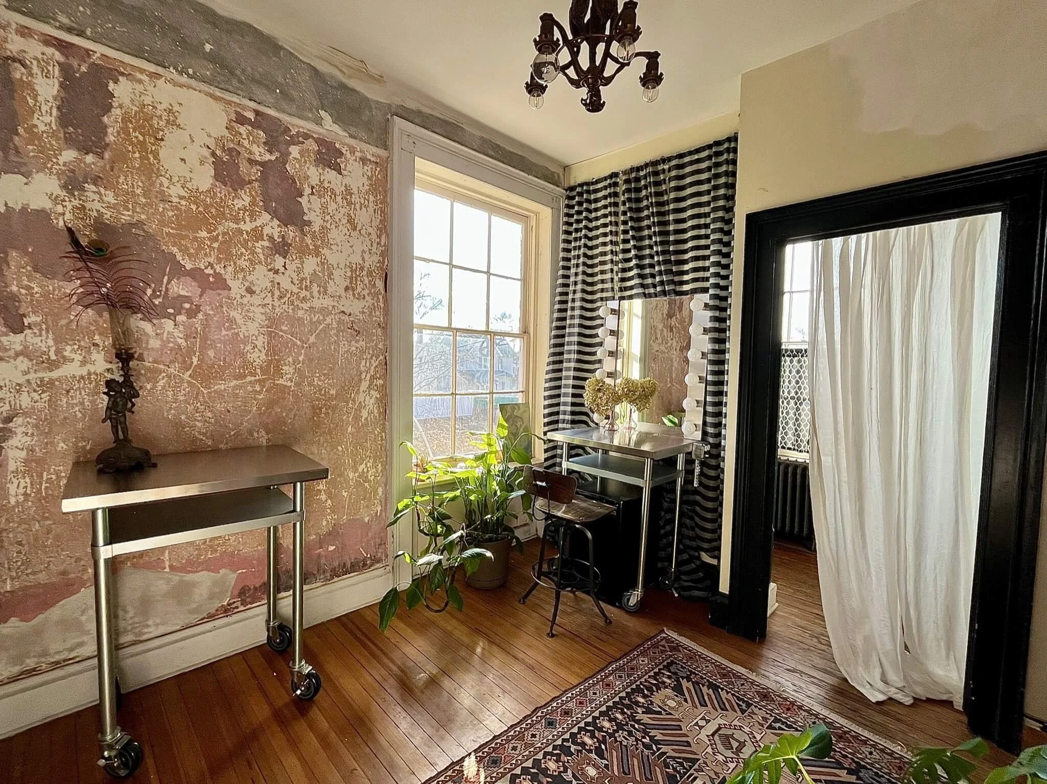 Interior of a room with a distressed wall, floral decor, a window with curtains, a dresser with a mirror and flowers, potted plants, and a patterned rug.