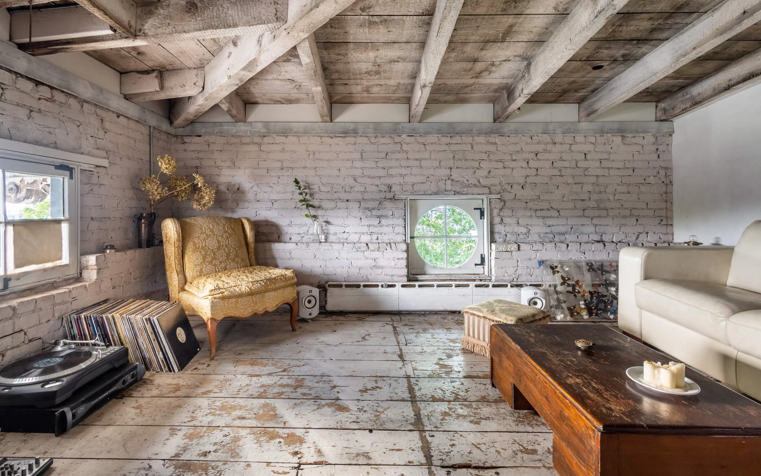 Living room with white brick walls and exposed wooden beams on the ceiling, featuring a vintage gold armchair, a white leather sofa, a wooden coffee table with candles, a vinyl record collection, a DJ turntable, and a small round window showing trees