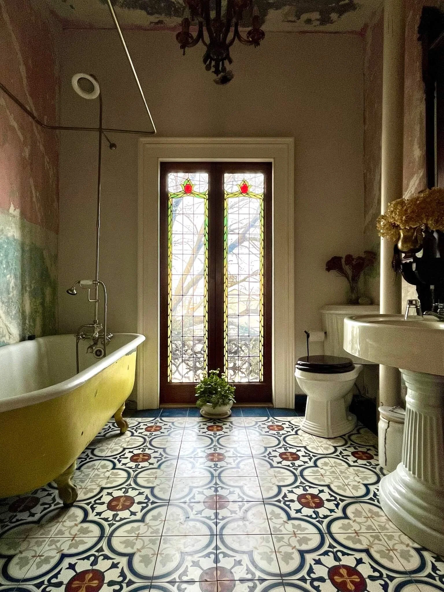 A vintage bathroom with a yellow clawfoot bathtub, stained glass door, patterned tile floor, toilet, and pedestal sink.