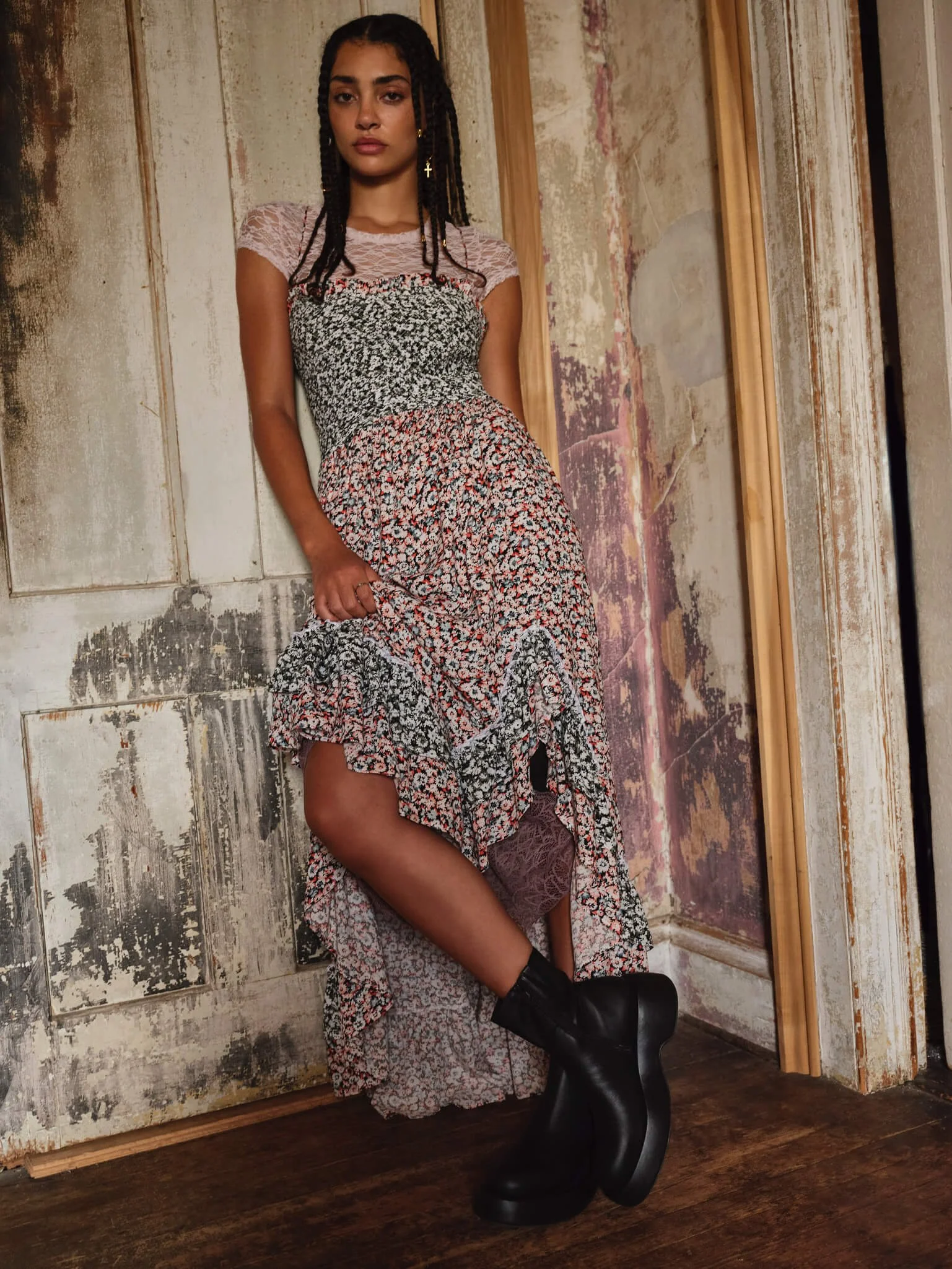 A woman with dark braided hair in a mixed floral dress with lace top, black platform boots, standing against a distressed vintage wall.