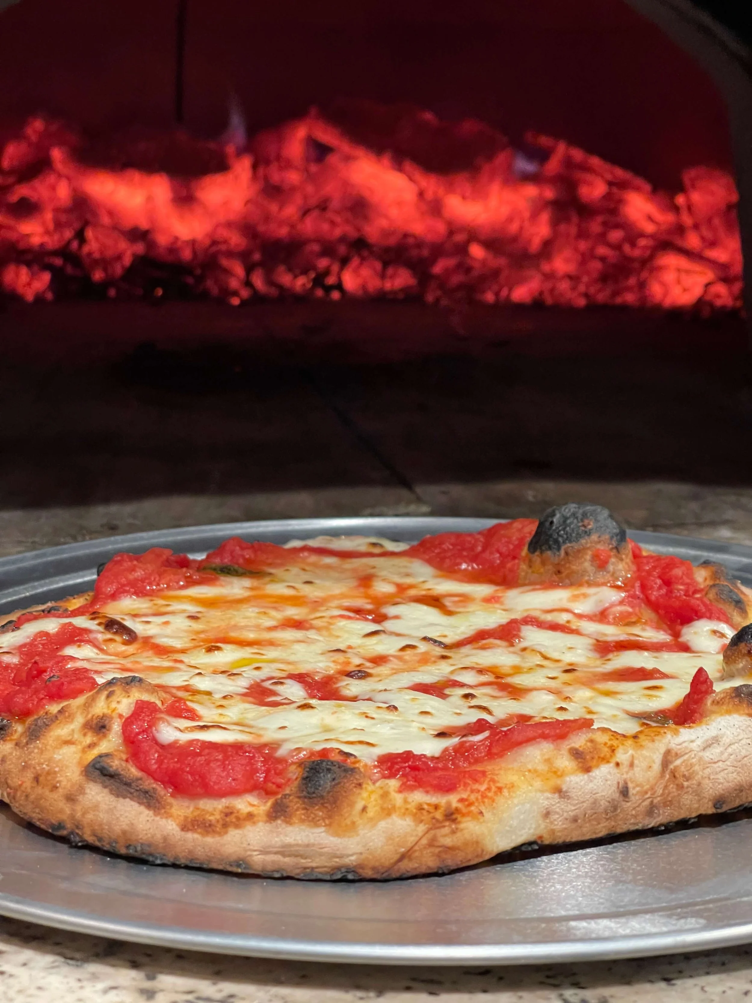 A cheese pizza with tomato sauce and a slightly burnt crust in front of a wood-fired oven with glowing flames inside.