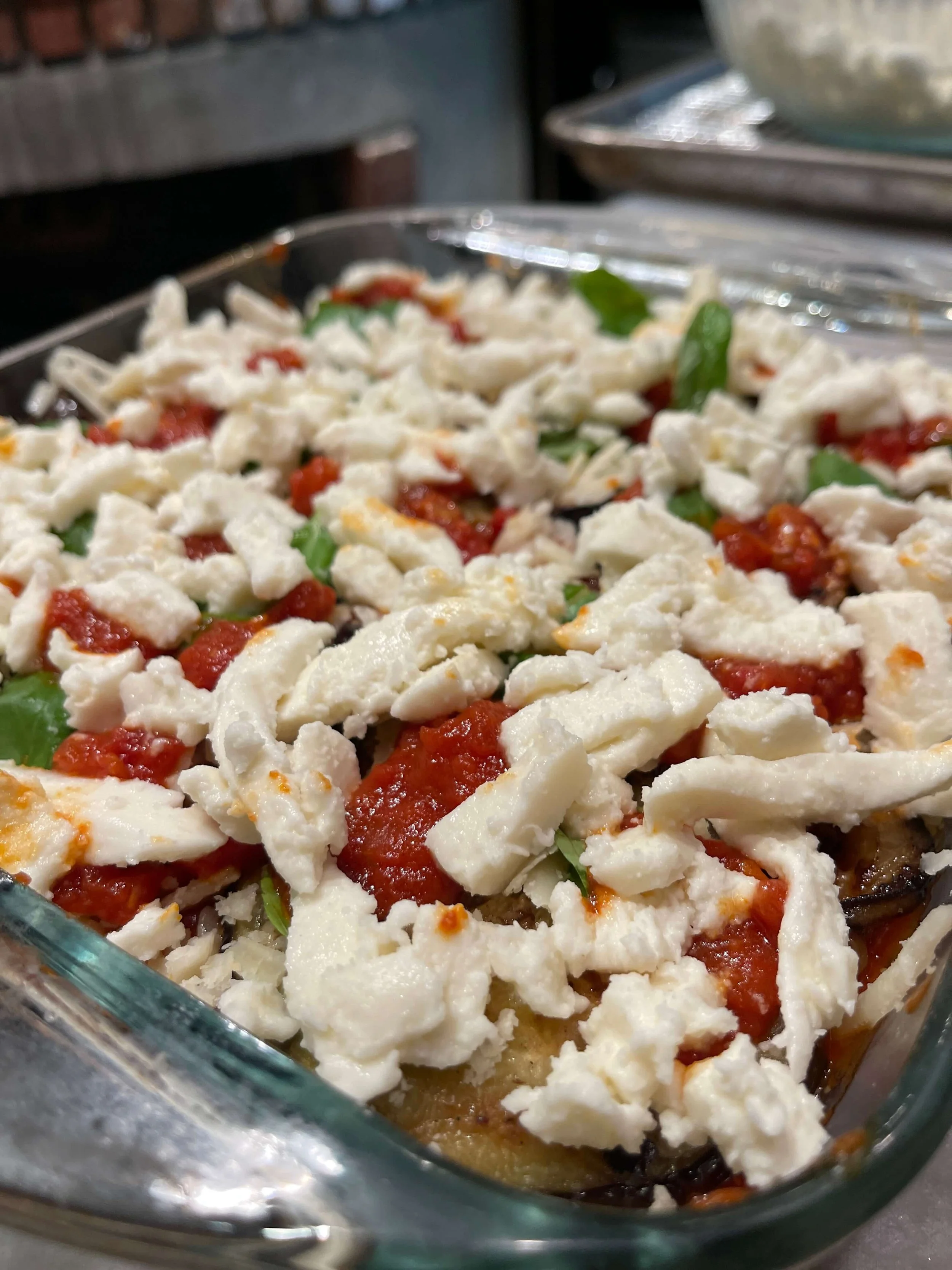 A close-up of a baked eggplant dish topped with chopped mozzarella cheese, tomato sauce, and basil leaves in a glass baking dish.