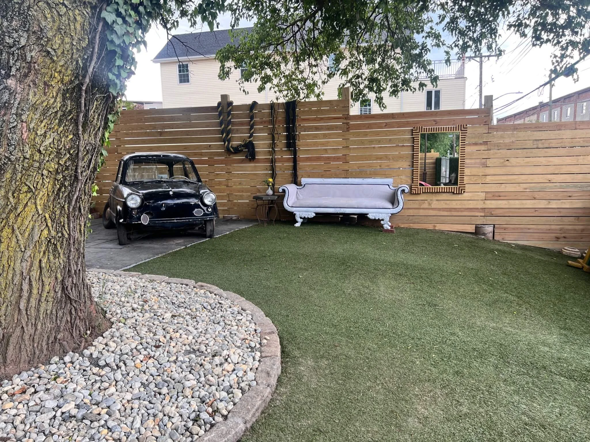 Backyard with artificial grass, a large tree, a pebble garden border, a vintage black car, a small side table with a flower vase, a white vintage-style sofa, a wooden fence with hanging ropes, a framed mirror on the fence, and a house with a balcony 