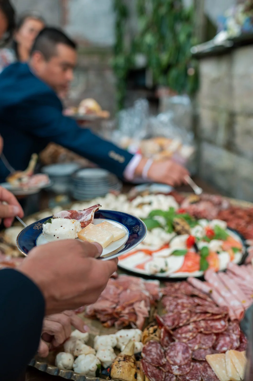 People serving themselves food at a buffet table with a variety of cheeses, meats, and vegetables, with blurred individuals in the background.