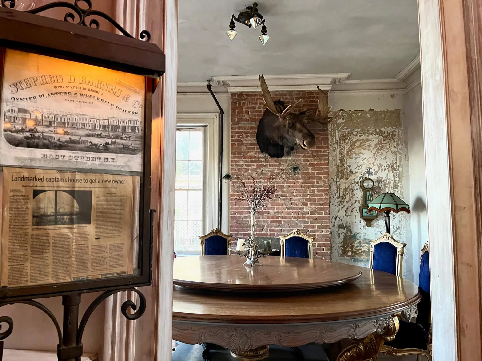 A vintage dining room with a large round wooden table and blue velvet chairs. A mounted deer head is on a brick wall, and a distressed wall with peeling paint. There's an antique lamp with a stained glass shade and a small clock. Sunlight comes throu