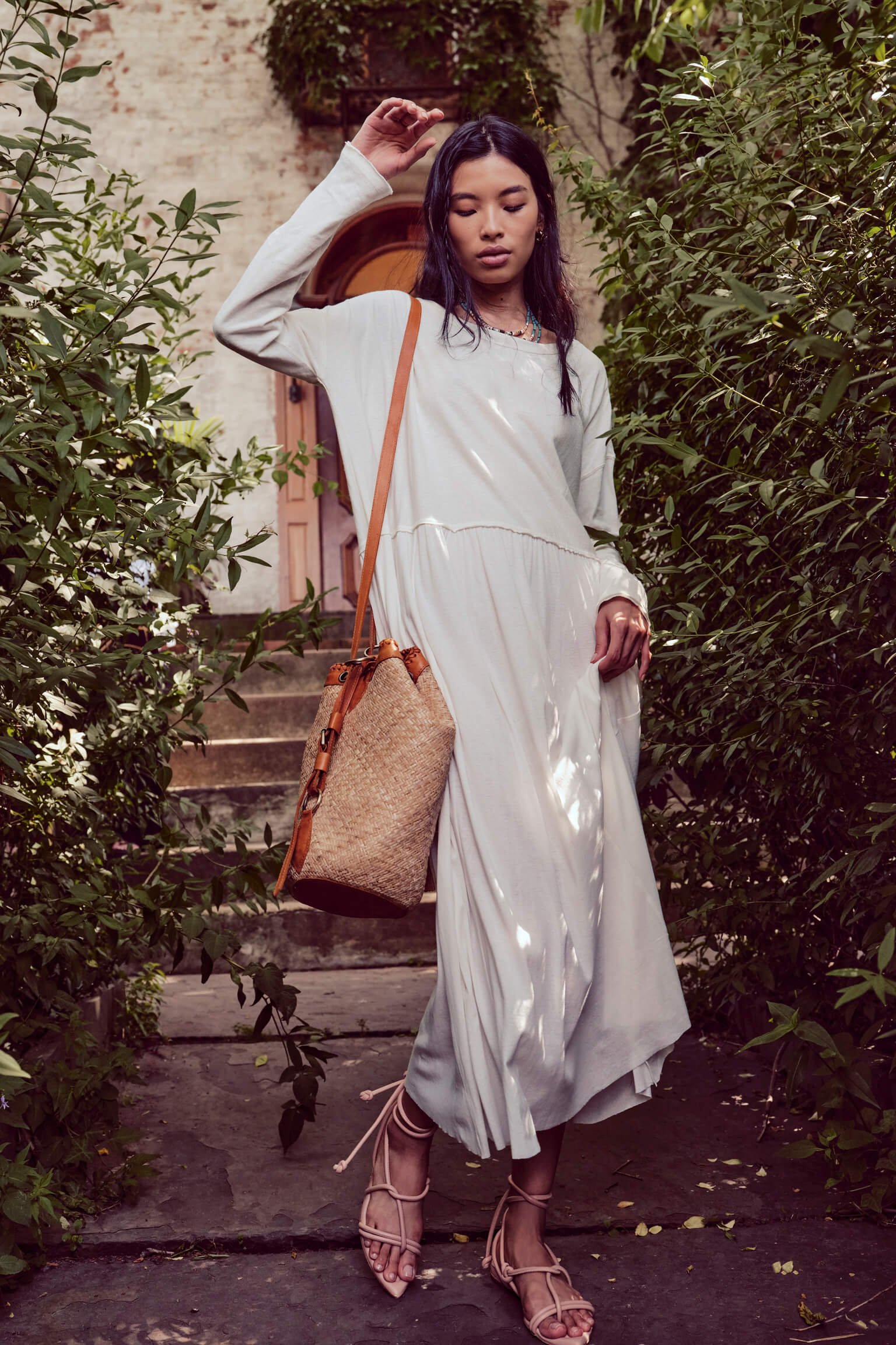 A young woman with long dark hair wearing a loose white dress, tan strappy heels, and a woven straw bag, standing on steps surrounded by lush green foliage near an arched doorway.