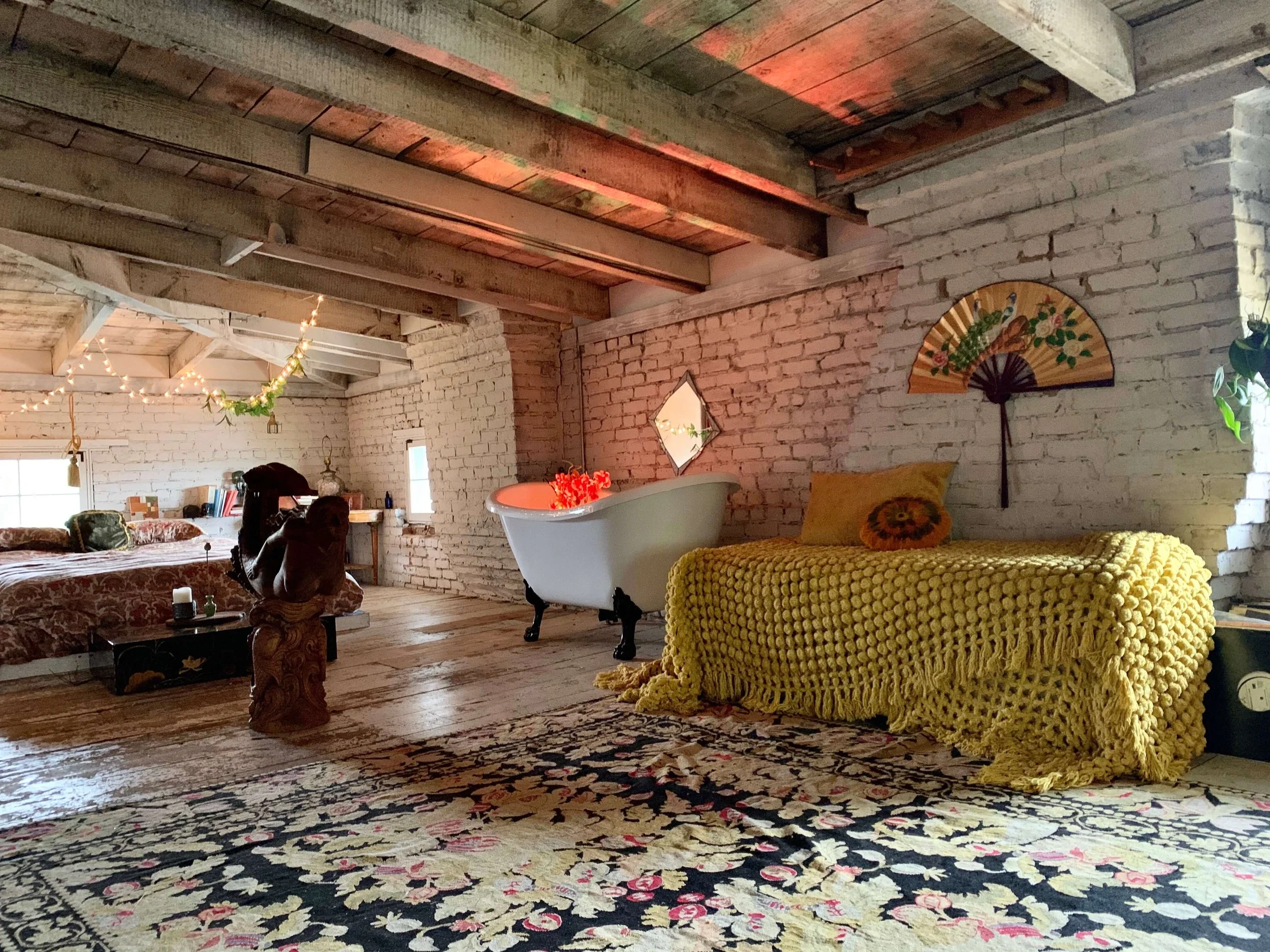 An eclectic loft with white brick walls, wooden ceiling beams, and a floral patterned rug. It features a white clawfoot bathtub, a yellow knitted sofa with a decorative pillow, and a bed with floral bedding. There are string lights, a fan-shaped wall