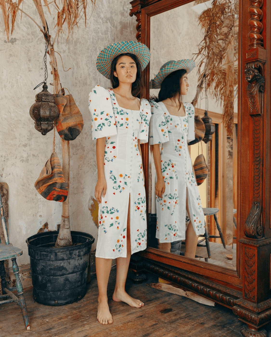 A woman wearing a white dress with colorful embroidery, a large straw hat, and barefoot standing next to a mirror in a rustic room with dried plants and hanging woven bags.