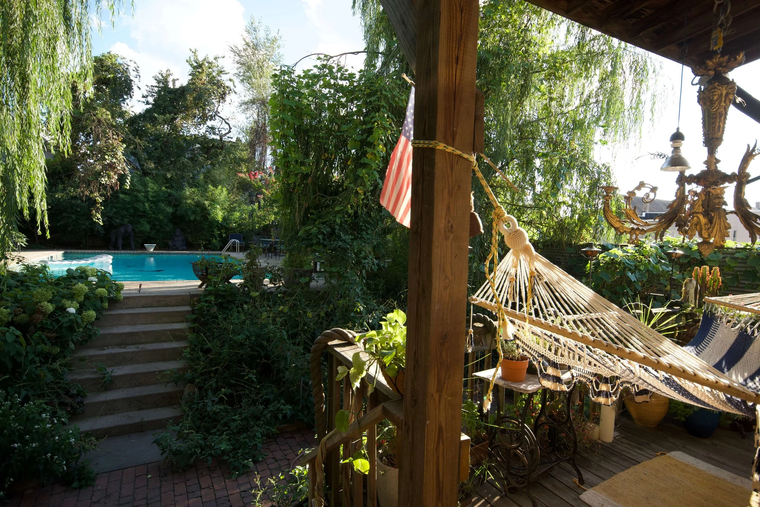A backyard patio with a hammock, potted plants, a chandelier, and a set of stairs leading to a swimming pool surrounded by lush greenery.