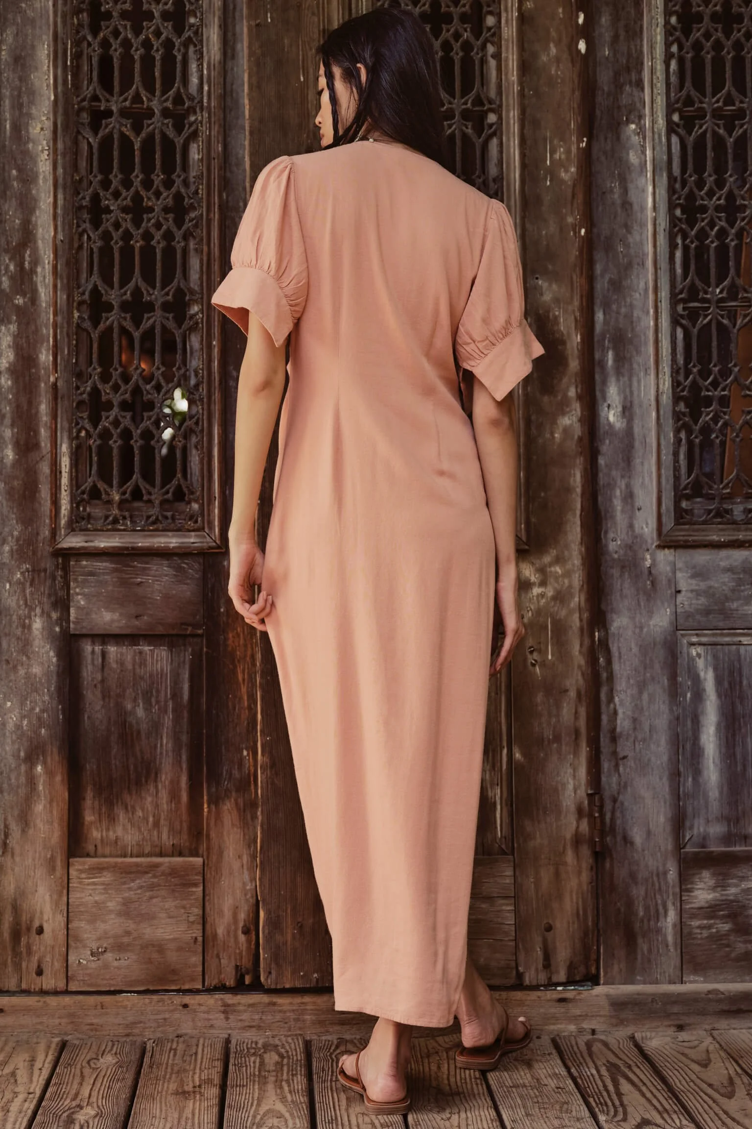 Woman in a pink dress standing with her back to the camera in front of rustic wooden doors.