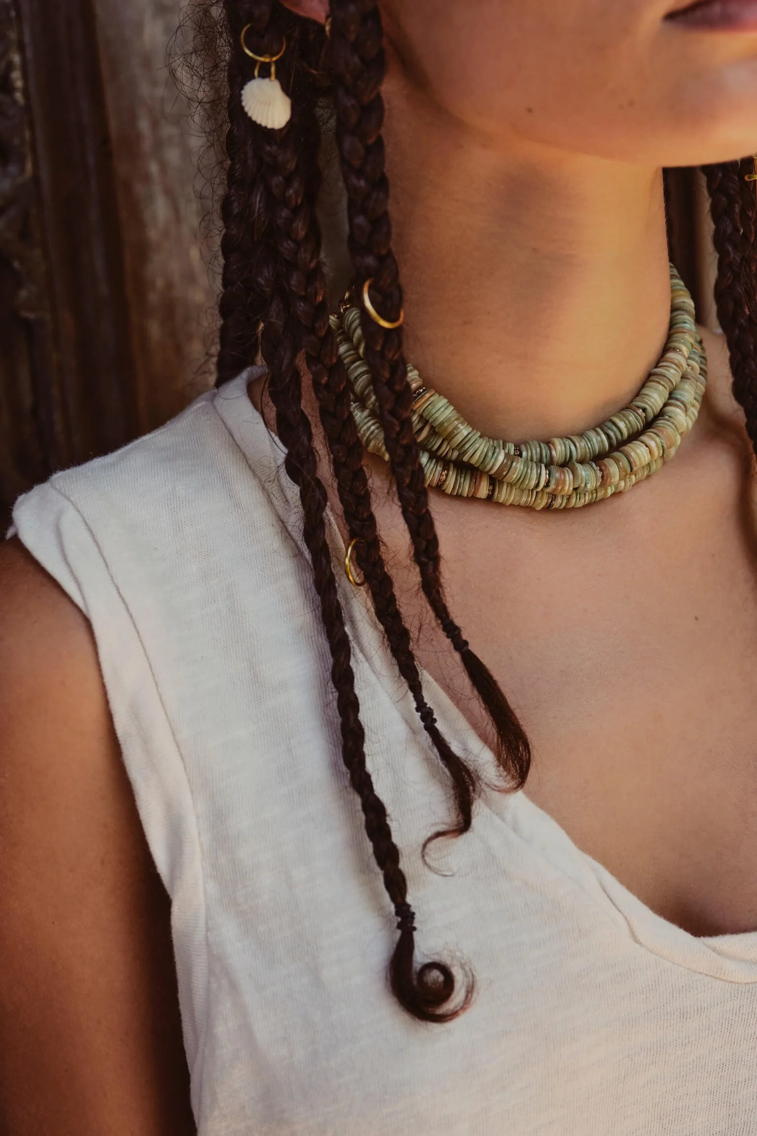 Close-up of a woman wearing layered necklaces and earrings, with braided dark hair and a white sleeveless top, against a wooden background.