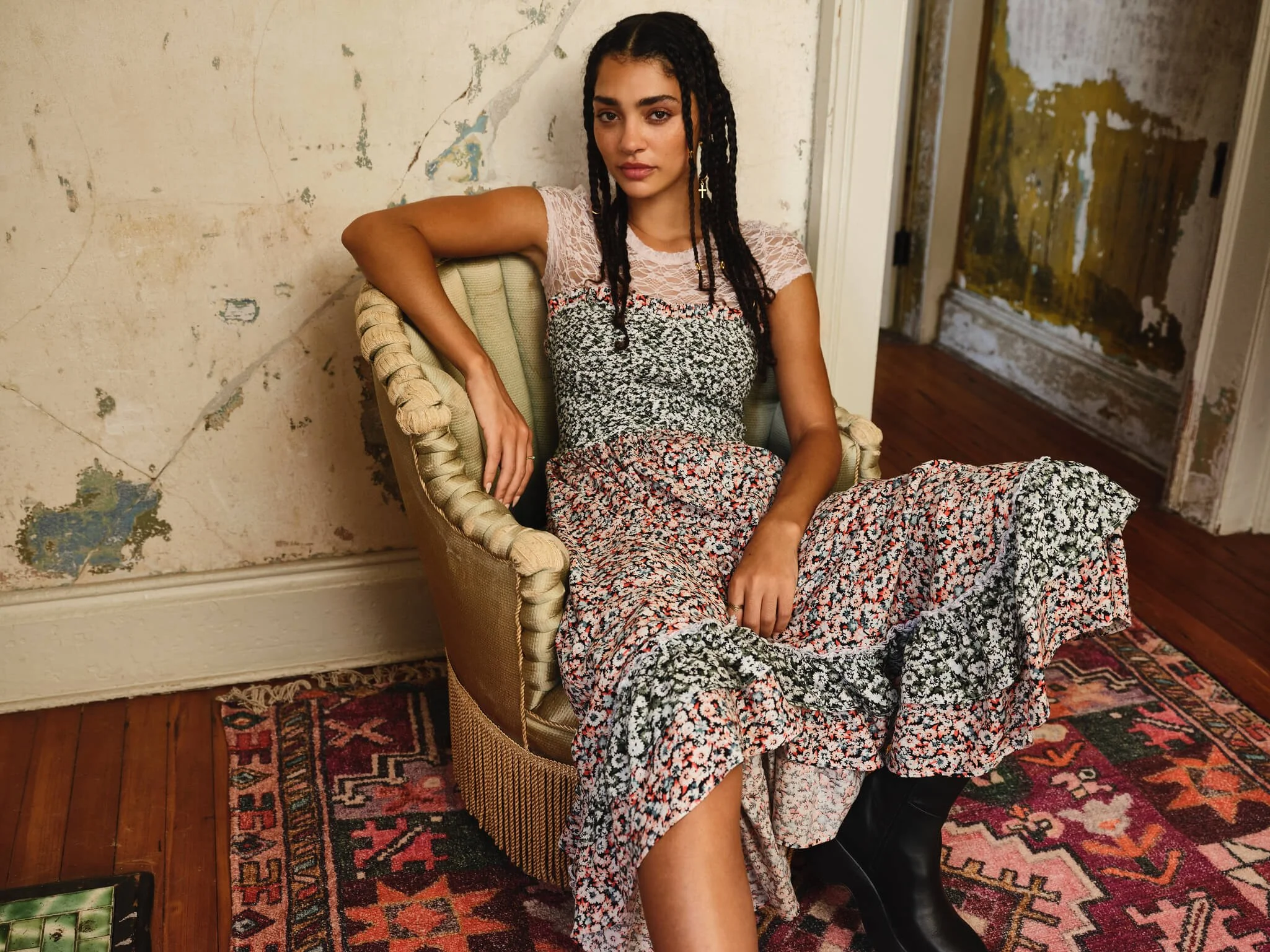 Young woman with braided hair sitting on a vintage armchair in a room with distressed walls and a patterned rug.