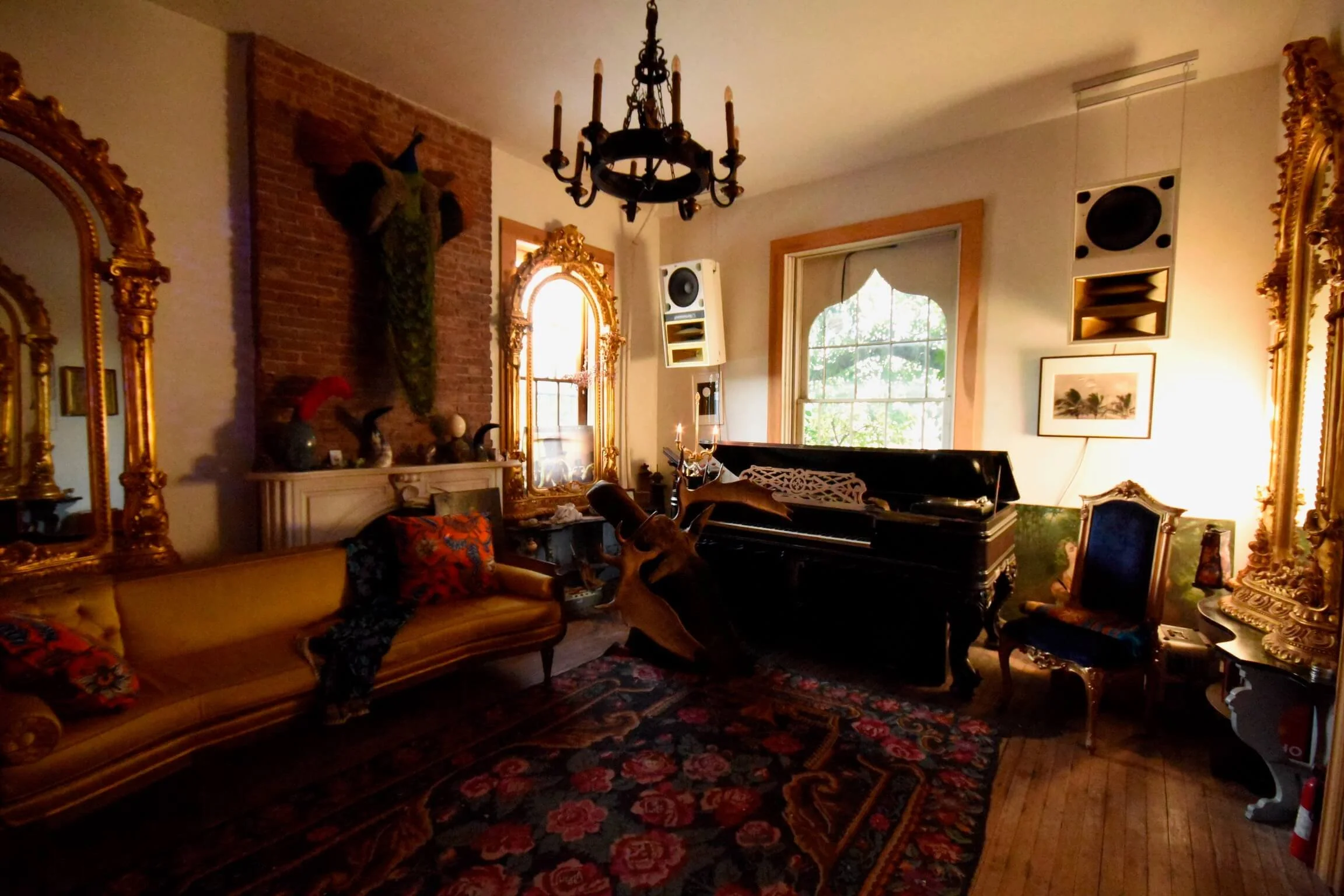 A living room with vintage and ornate decor, including large gold-framed mirrors, a black grand piano with a mounted deer head, a yellow velvet sofa with colorful cushions, a patterned area rug, and a chandelier hanging from the ceiling. There is a l