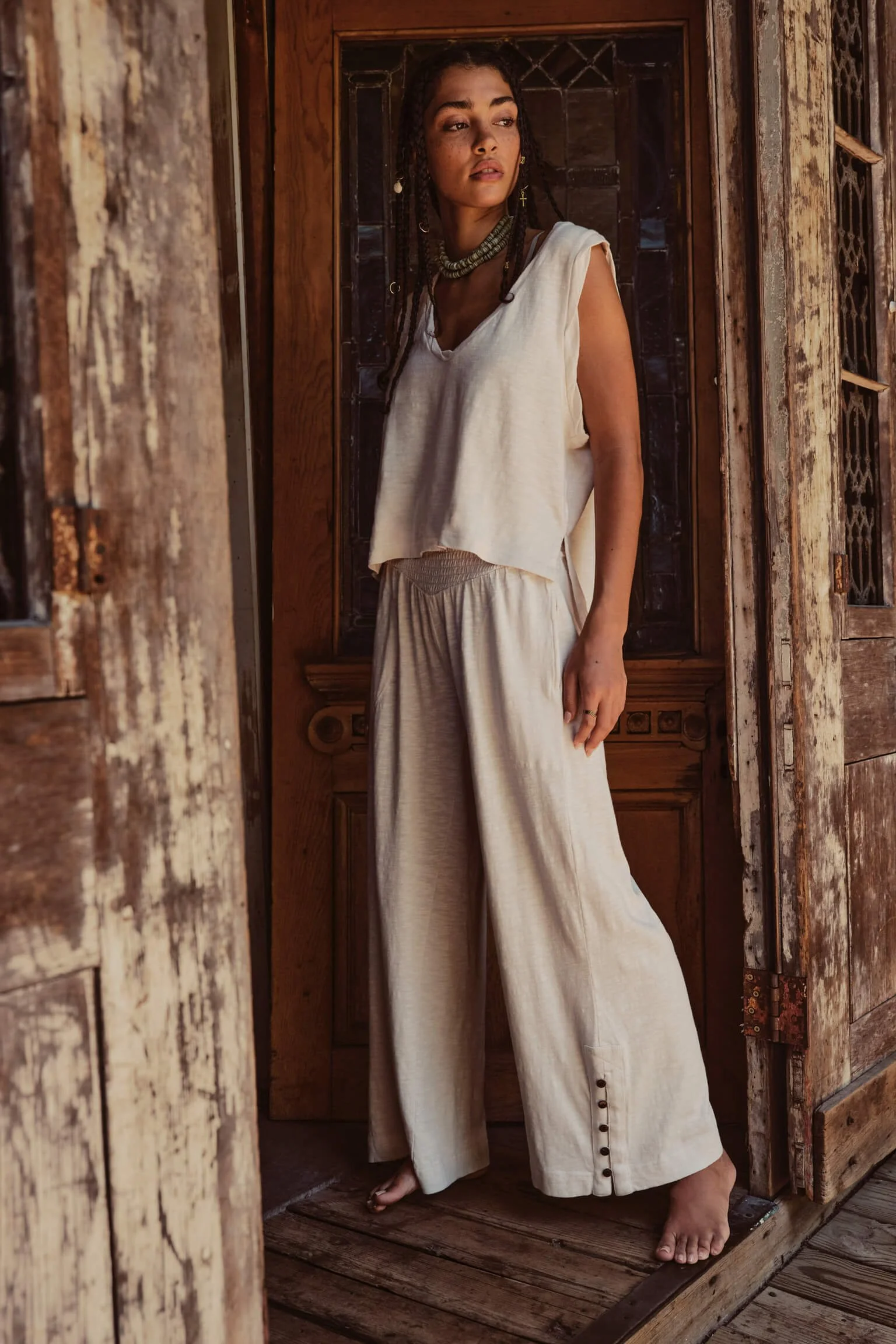 A woman with dark hair styled in braids, wearing a sleeveless cream top and matching wide-leg pants, standing barefoot in a rustic wooden doorway with weathered wood and stained glass.