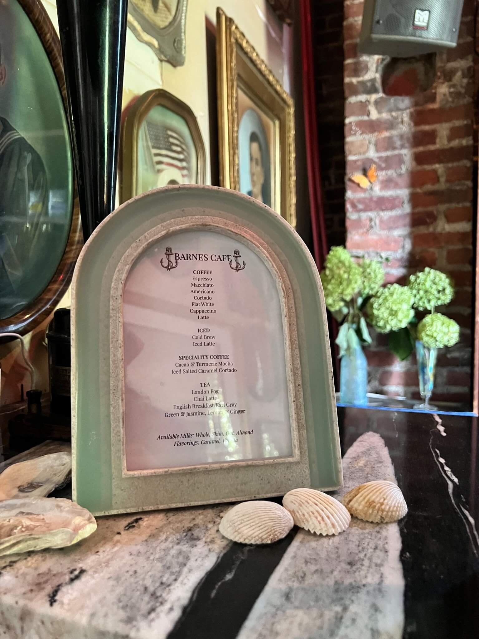 A menu from Barnes Cafe displaying coffee, iced coffee, specialty drinks, and tea options, with a rustic decor featuring framed pictures, shells, and a brick wall.