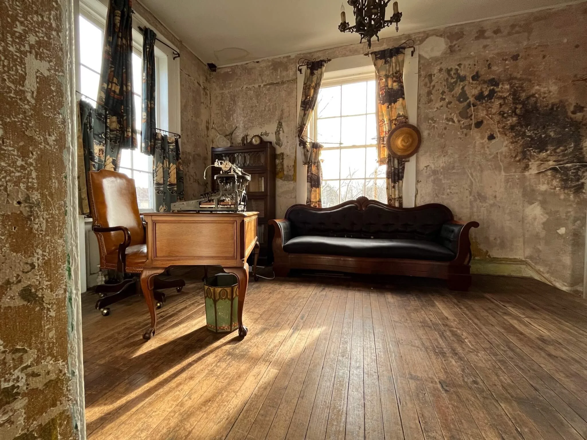 An old room with peeling wallpaper, wooden floors, a vintage desk and chair, a black leather sofa, and a window with patterned curtains letting in natural light.