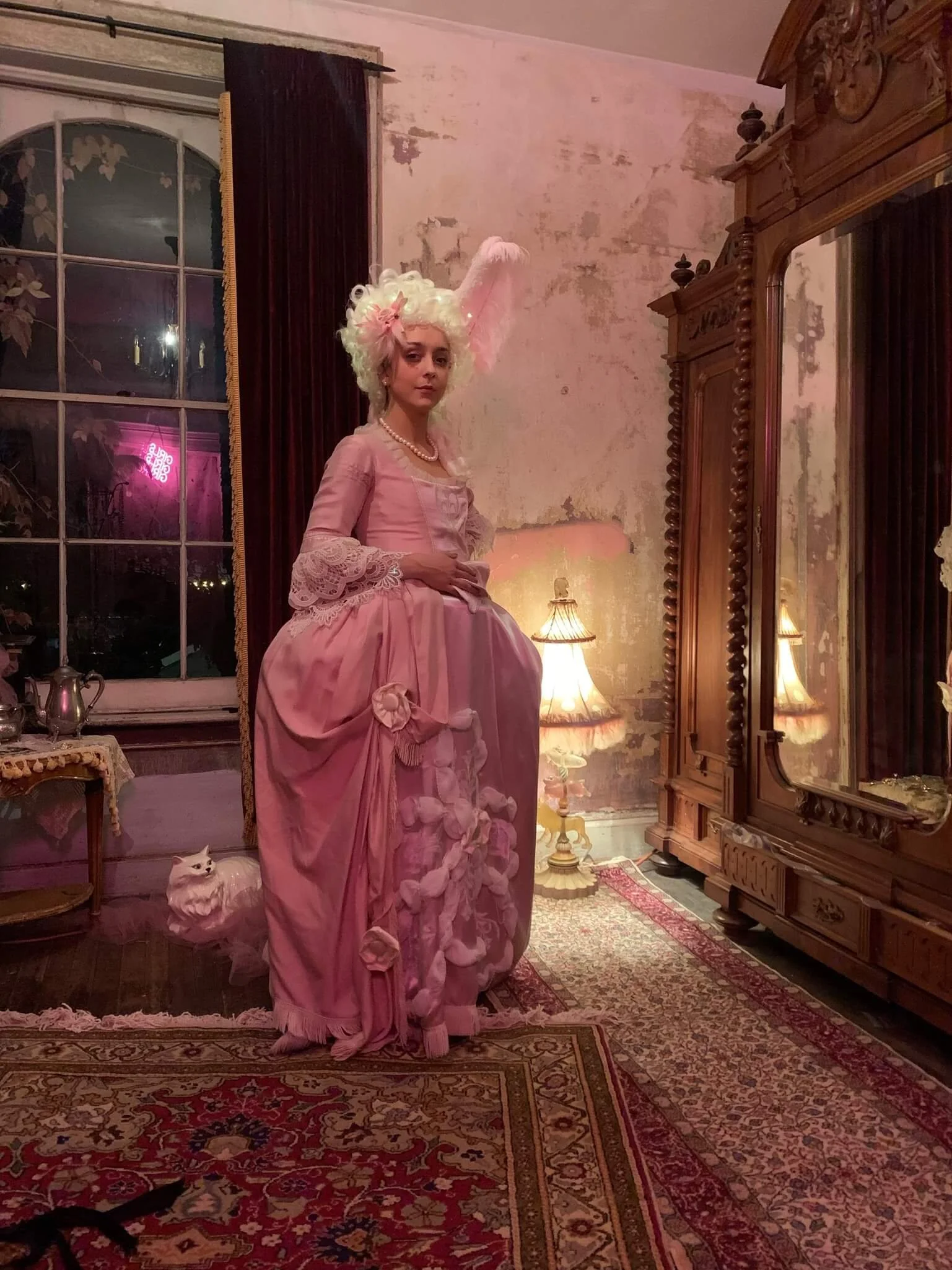 A woman dressed in an elaborate pink 18th-century style gown with lace sleeves and floral decorations, posing next to an antique wooden wardrobe in a vintage-styled room with peeling wall paint, a patterned rug, and warm table lamps.