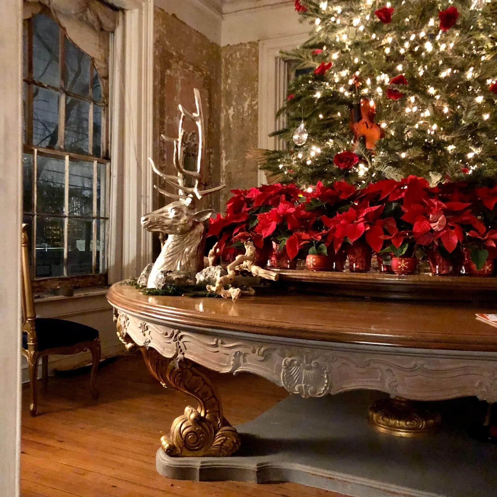 A decorated Christmas tree with lights, ornaments, and red ribbons, placed on a table with poinsettia flowers. In front of the table, there is a decorative reindeer sculpture, and the room has a rustic vibe with exposed brick walls and wooden floor.