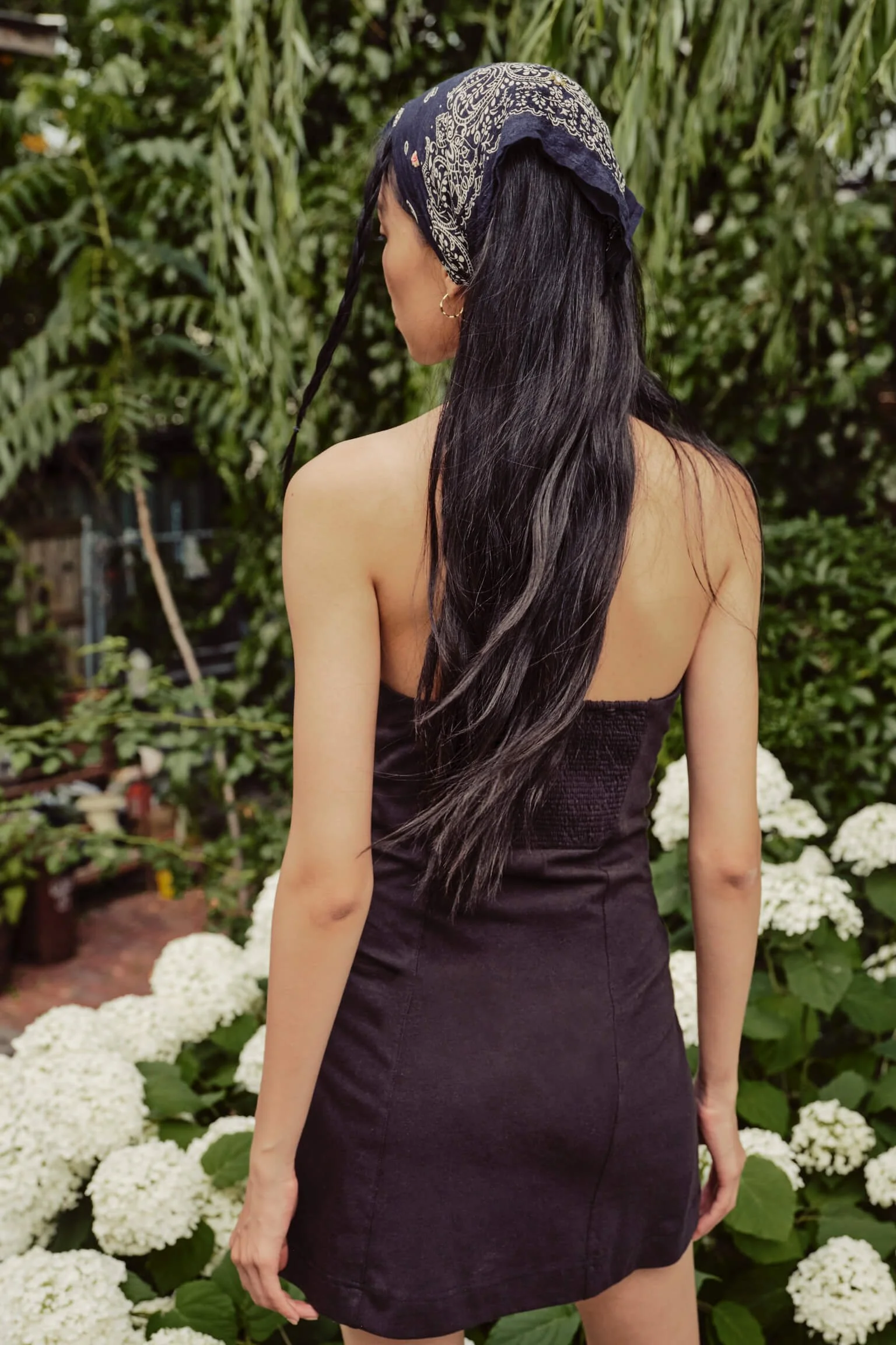 A woman with long black hair wearing a black dress and a bandana is standing in front of white hydrangea flowers and green foliage.