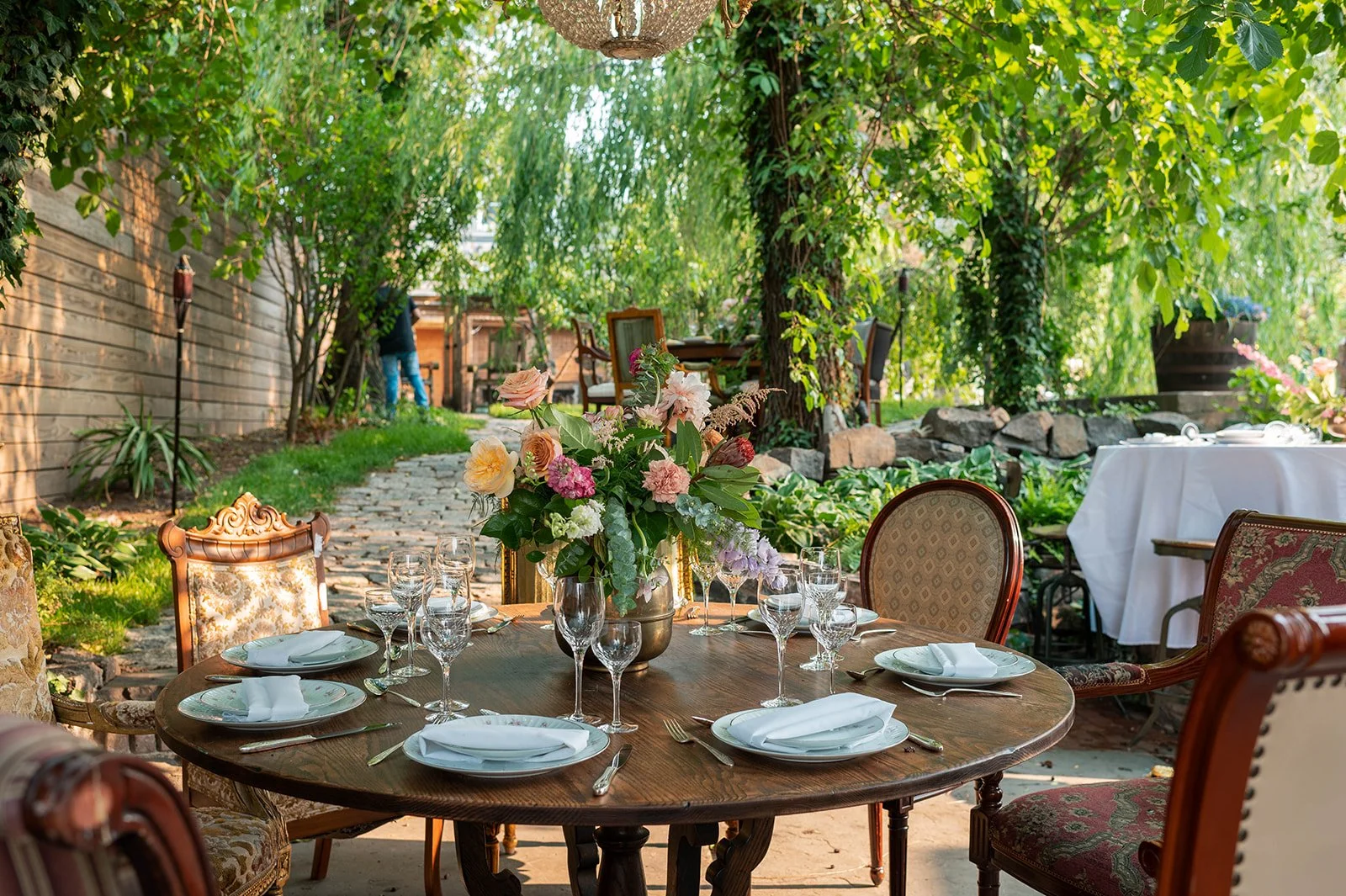 Elegant outdoor dining setup with a round wooden table, floral centerpiece, white napkins, glassware, and vintage chairs, in a lush garden with green trees and a stone pathway.