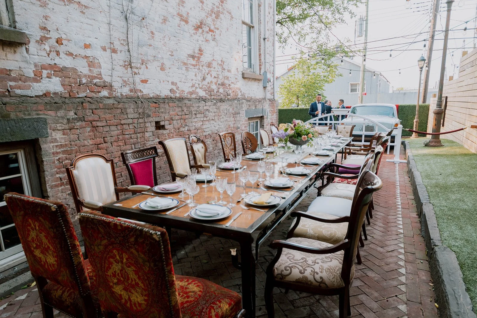 Elegant outdoor dinner party setup with a long wooden table, decorated with a floral centerpiece, surrounded by vintage upholstered chairs on a brick-paved patio. In the background, a brick wall, a vintage car, and a few people in suits chatting