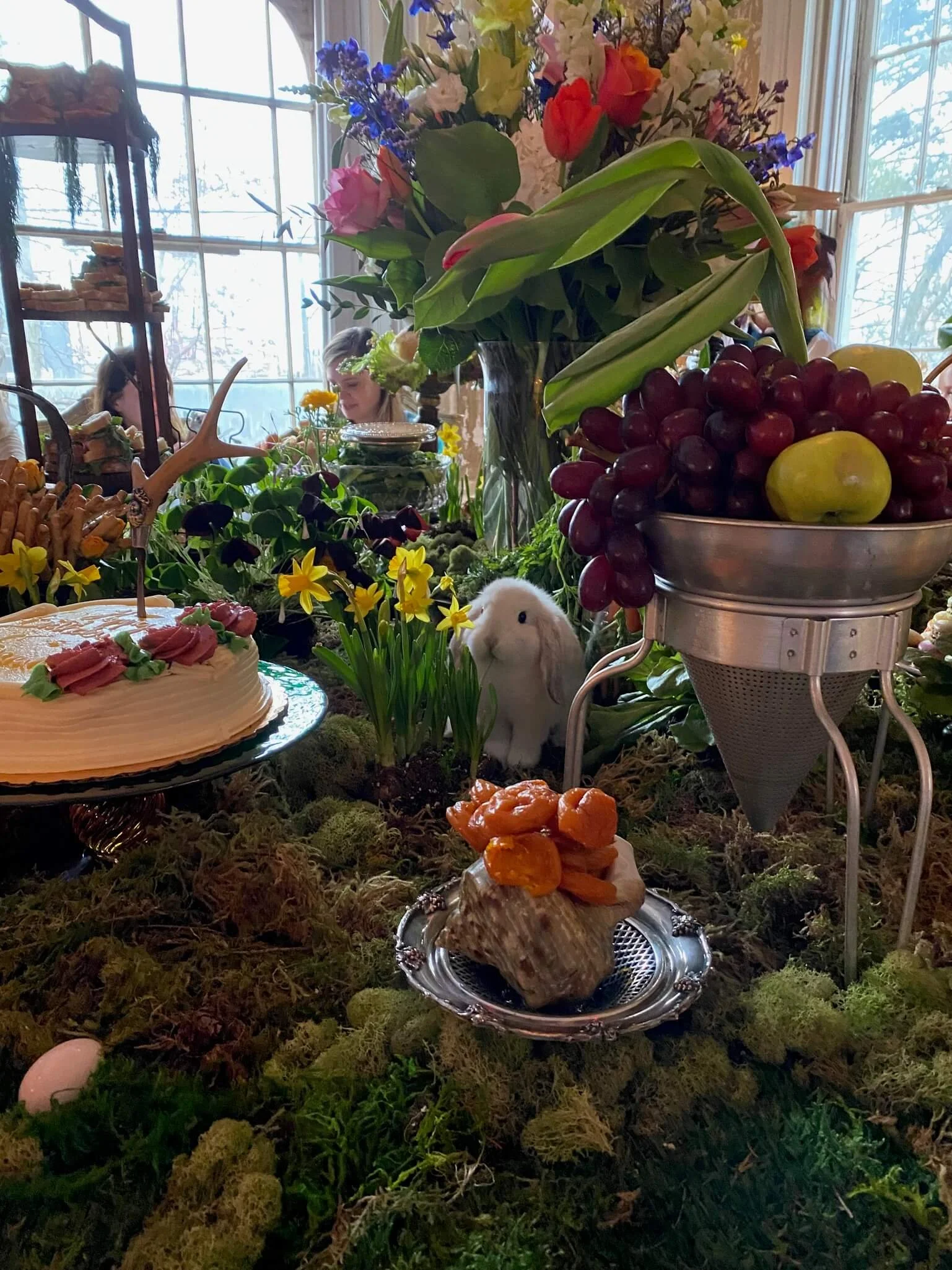 A table filled with fruits, flowers, a cake, a chocolate-covered treat with orange toppings, and a white rabbit figurine. In the background, a large window shows a bright outdoor scene, with some people visible inside near the window.