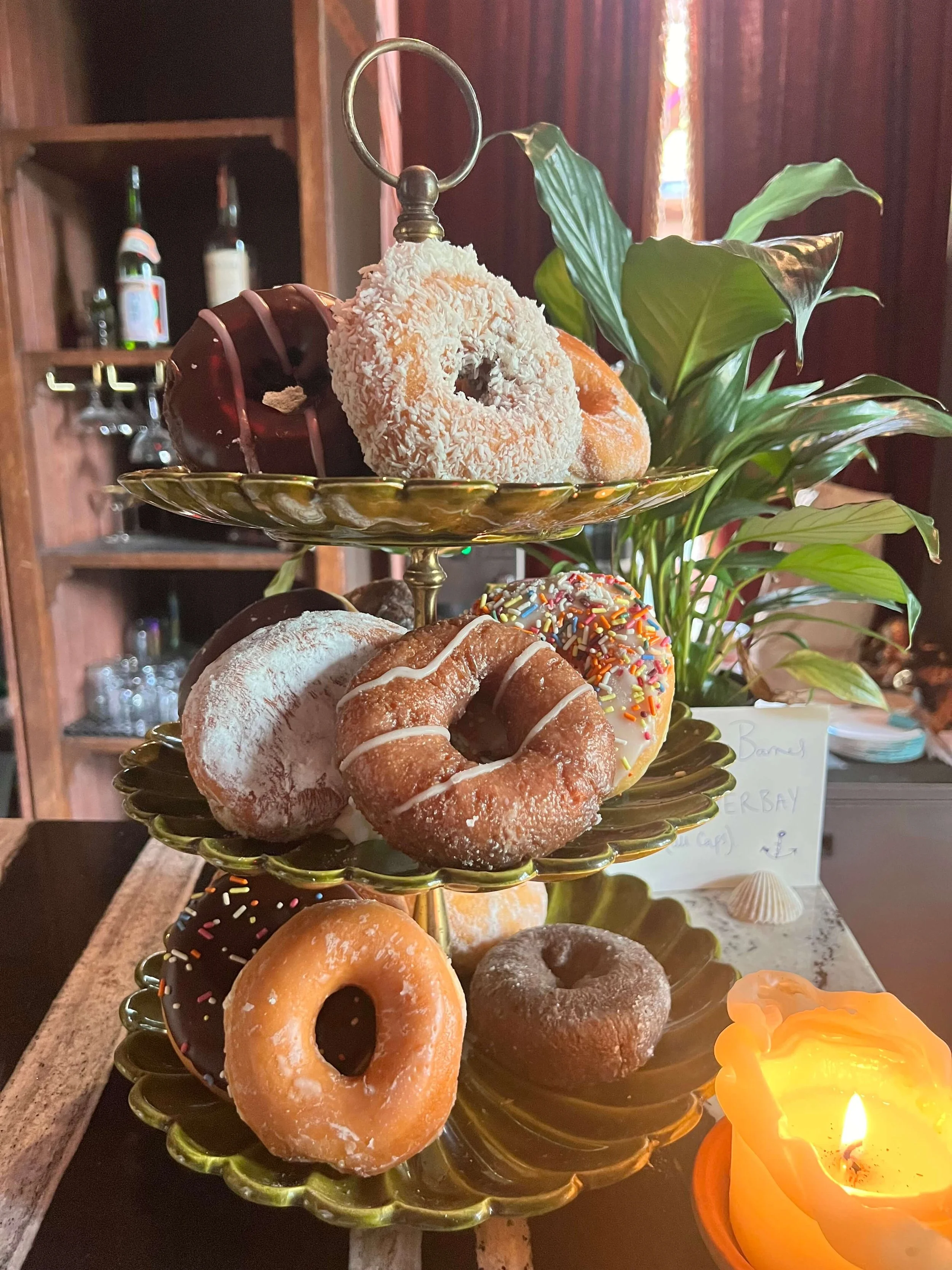 A three-tiered stand filled with assorted donuts of various flavors and toppings, placed on a table with a lit candle nearby and a plant in the background.