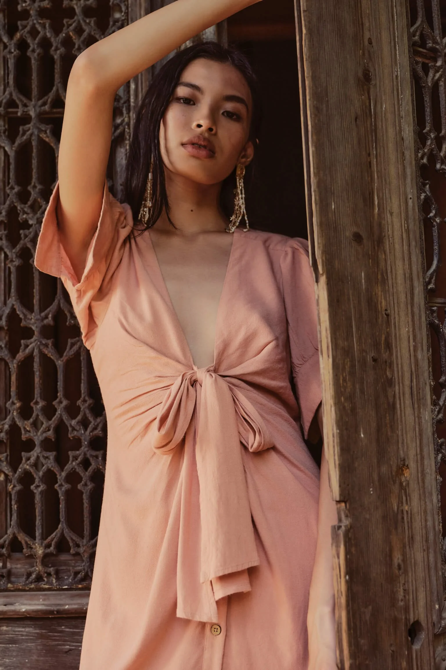 A woman with long dark hair and gold jewelry posing in a peach-colored dress with a deep V neckline and bow detail, standing next to a wooden post with a decorative grate in the background.