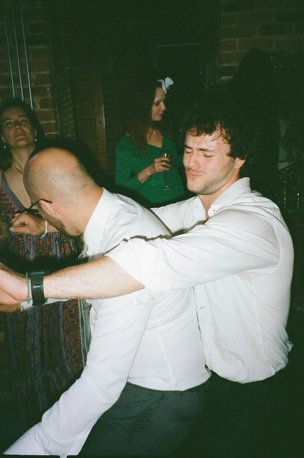 A man in a white shirt appears to be dancing or embracing a man with a bald head and glasses at a party or social gathering.