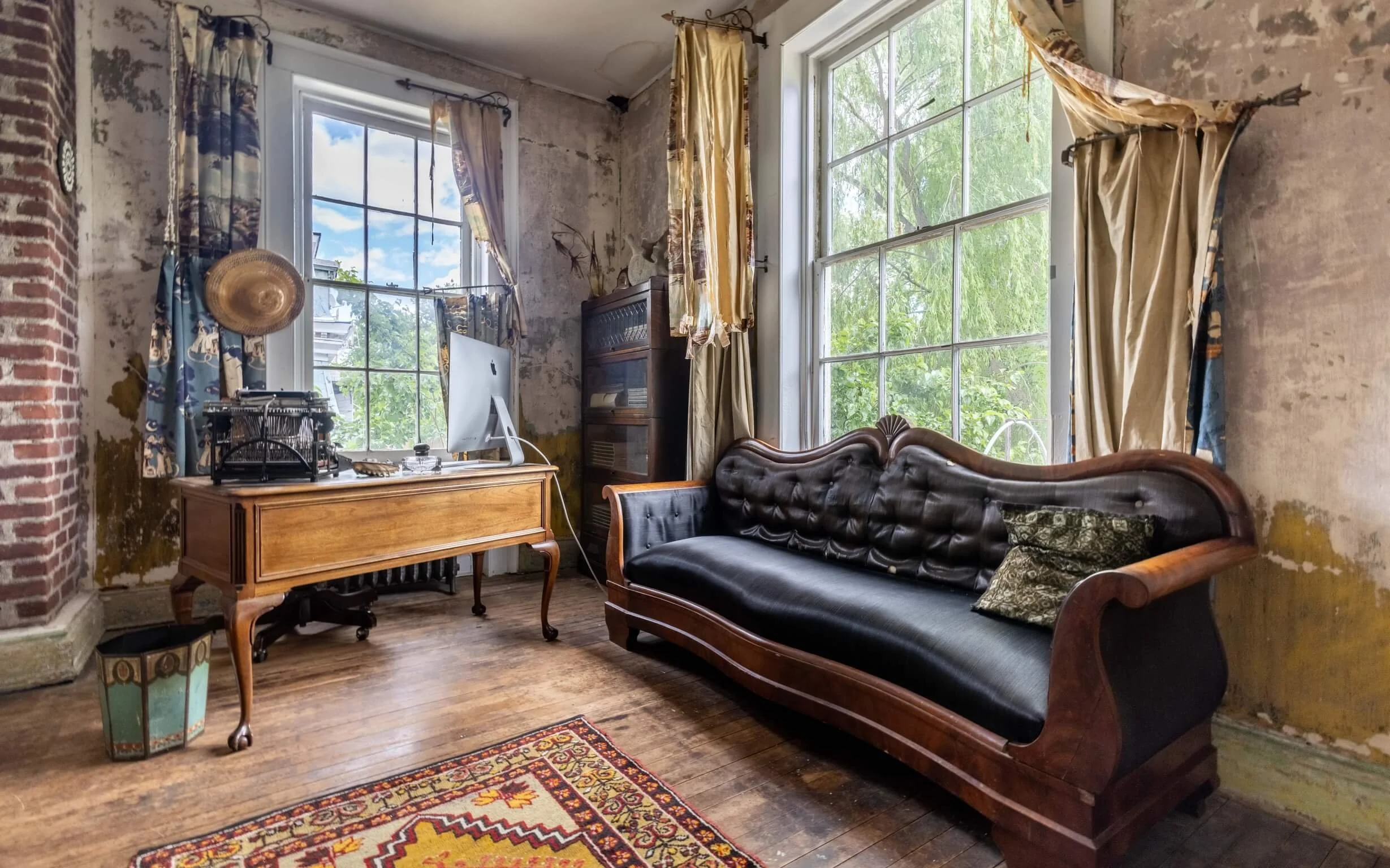 Vintage room with distressed walls, wooden flooring, large windows with curtains, a black leather and wood sofa, a wooden desk with a computer, and a colorful rug.