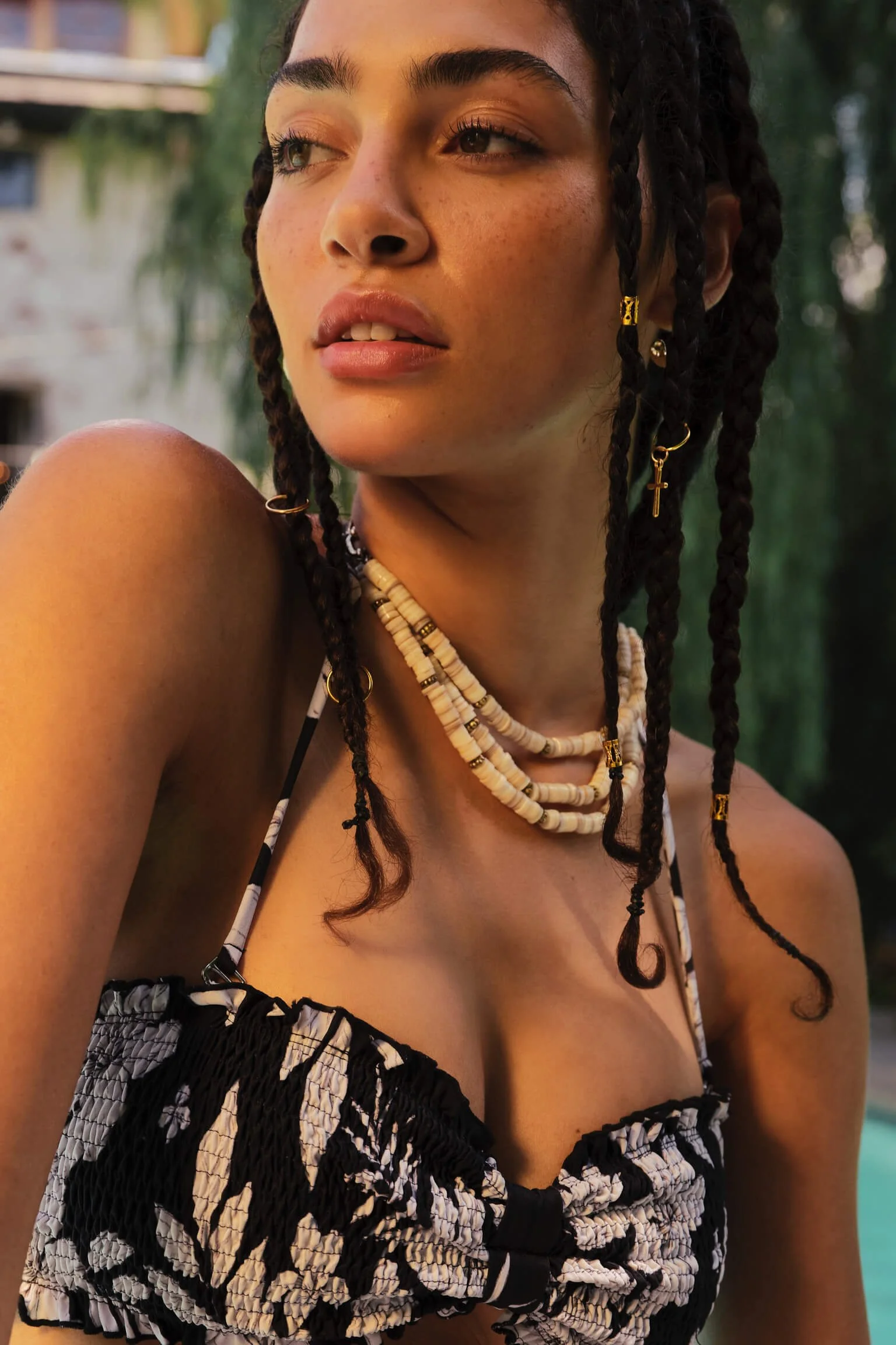 A woman with braided hair wearing layered necklaces, patterned swimsuit, and gold jewelry, looking to the side with a background of greenery and a building.