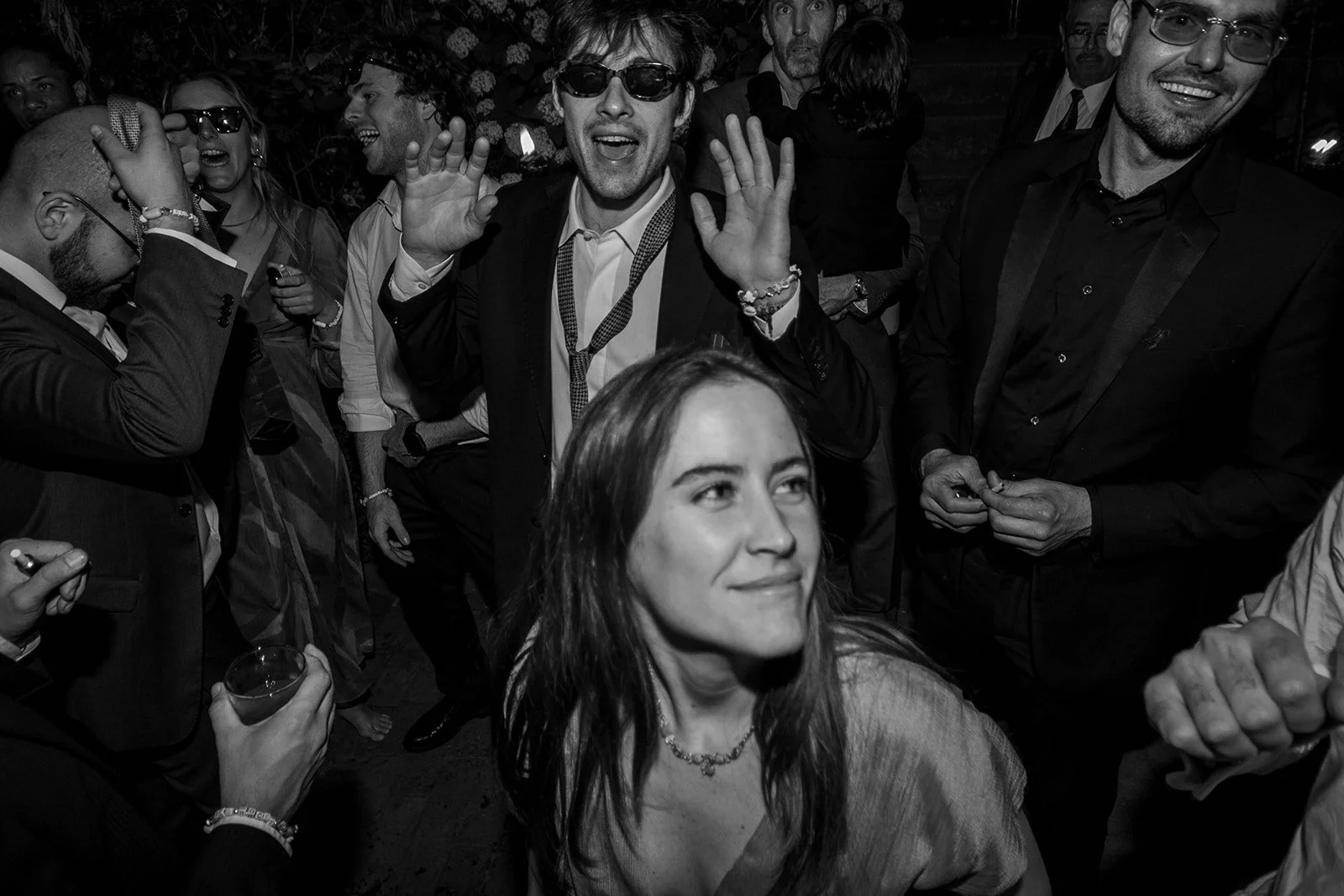 Black and white photo of a group of people celebrating at a party, with a woman in the foreground smiling and men in suits behind her, some dancing and some drinking.