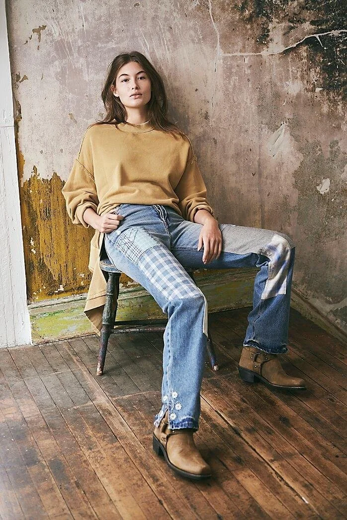 Young woman sitting on a vintage wooden bench in a rustic, distressed room with exposed walls and wooden floors. She wears a loose tan sweatshirt, patchwork jeans with floral embroidery, and tan ankle boots.