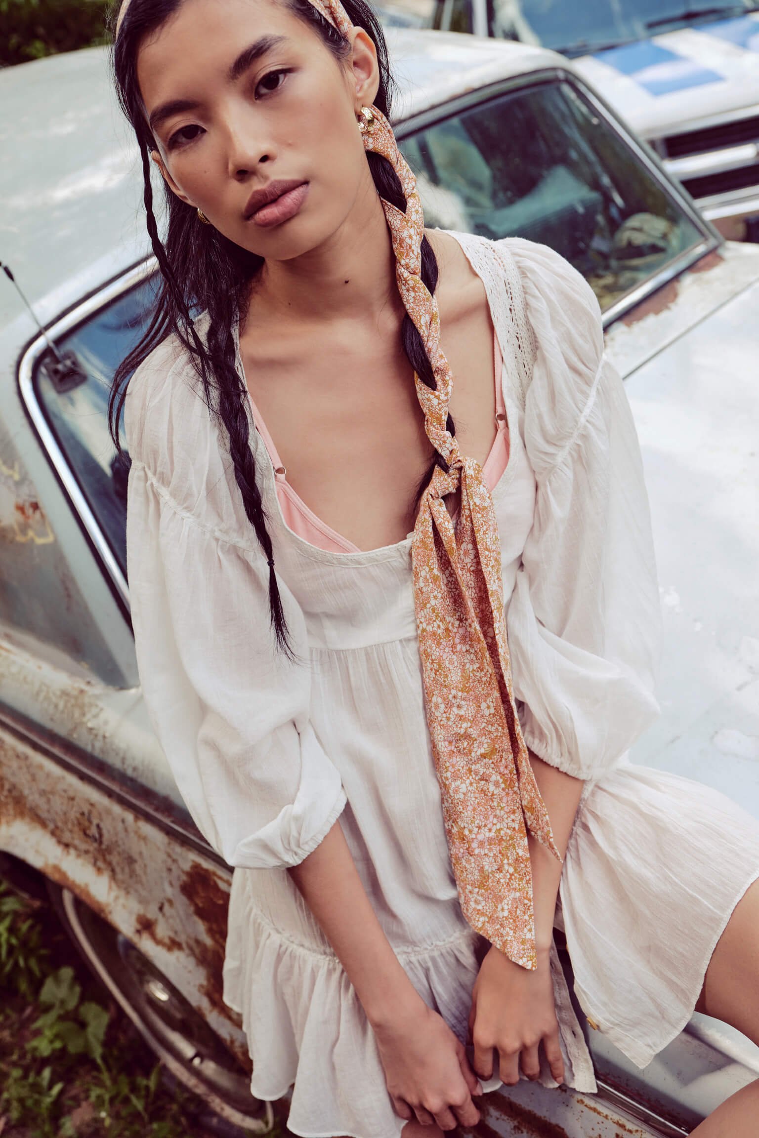 A young woman with dark hair and light skin, wearing a white dress with puffed sleeves and a pink camisole, standing next to a rusty vintage car. She has two braids and an orange floral scarf tied loosely around her neck.