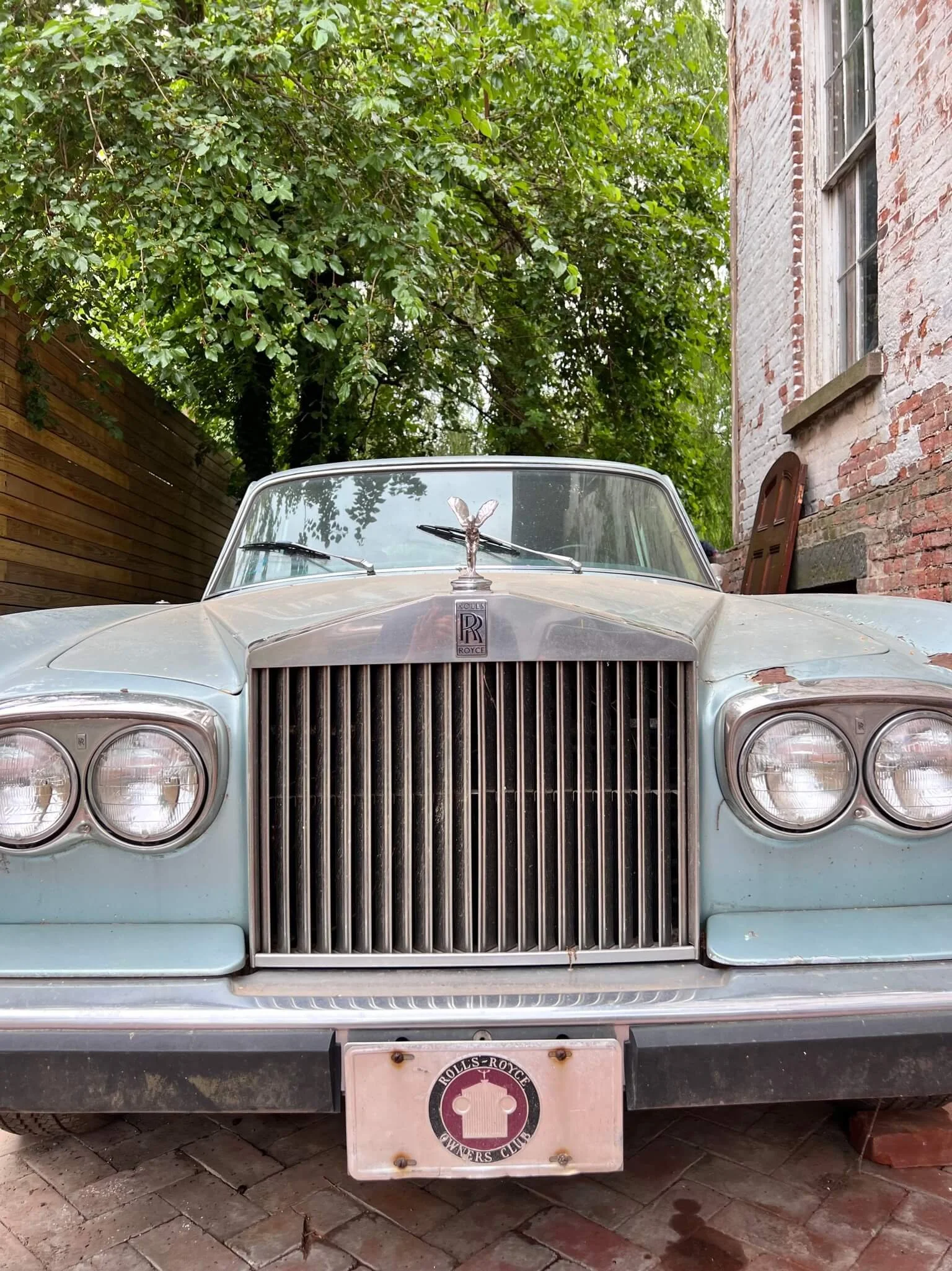 Front view of a vintage blue Rolls-Royce car with a hood ornament, parked on brick pavement beside a brick building and a wooden fence, surrounded by green trees.