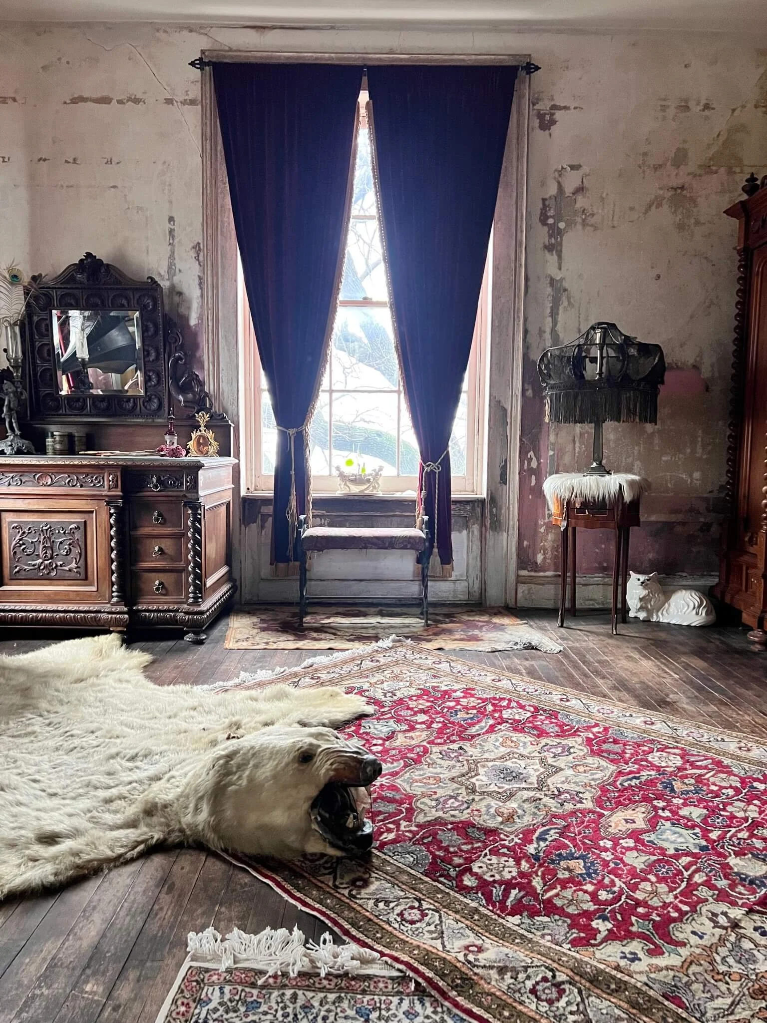 An antique-style room with a large window and dark purple curtains, an ornate wooden dresser, a fur rug with a bear head design, patterned rugs, and a small white ceramic dog statue.