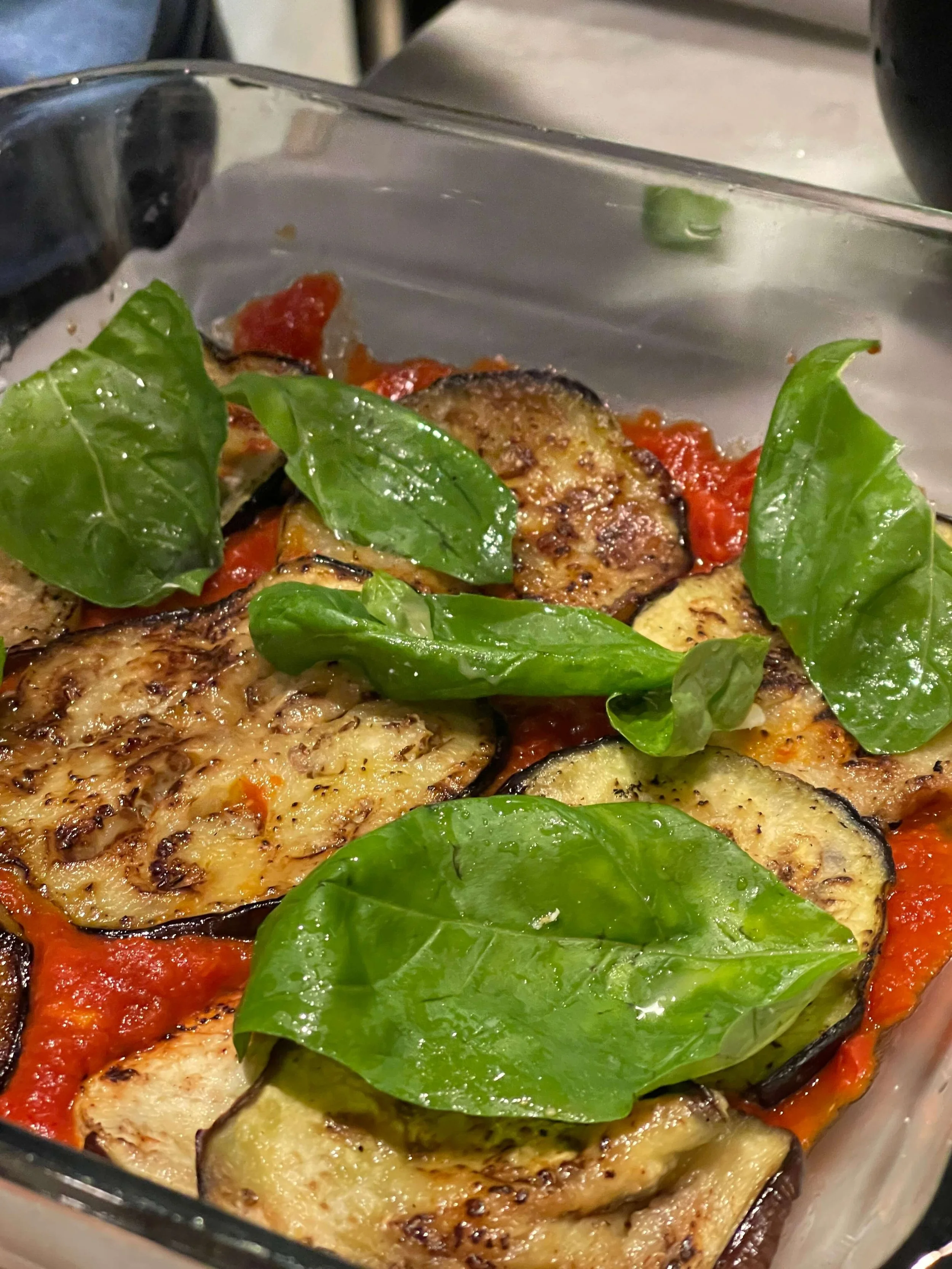 Baked eggplant slices with tomato sauce and fresh basil leaves in a glass dish.