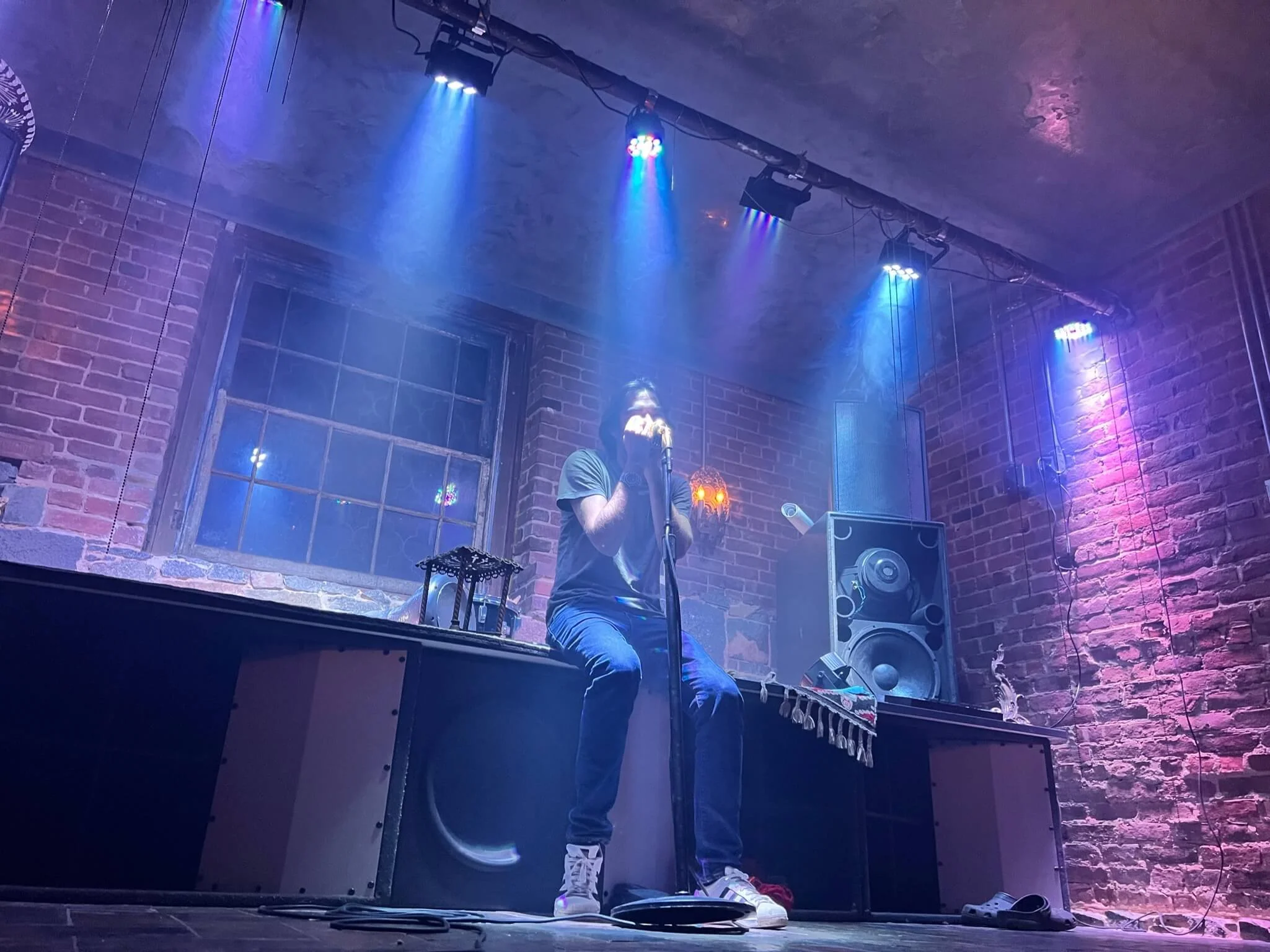 A person is sitting on a stage in front of a microphone, performing or speaking. The stage has brick walls and colorful lights overhead. There is a large speaker and some electronic equipment nearby.