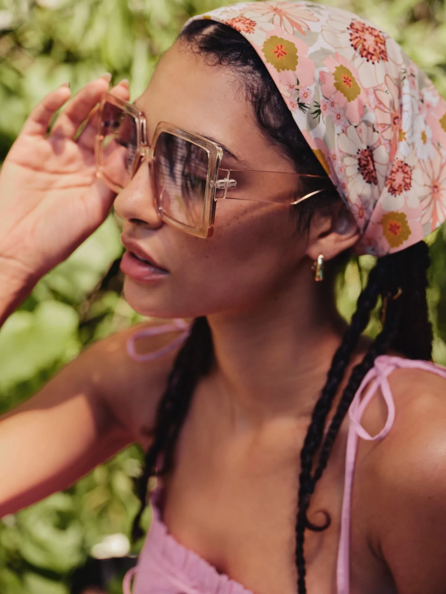 A woman with dark hair styled in braids, wearing large pink sunglasses, a pink floral headscarf, and a pink dress with thin straps, outdoors surrounded by green foliage.