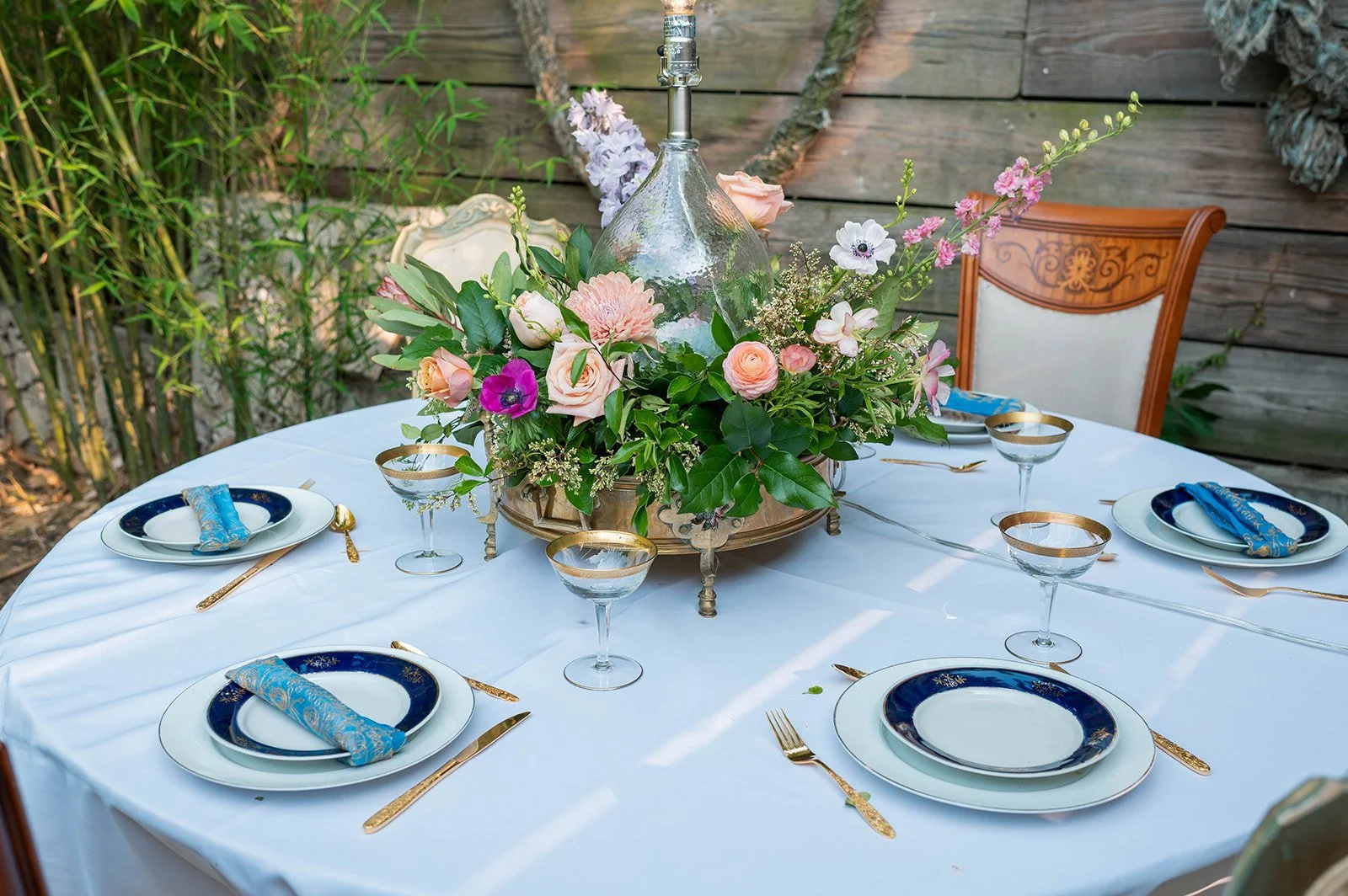 A round outdoor table set for four, with a white tablecloth, featuring a floral centerpiece with various pink, white, and purple flowers in a large glass vase, surrounded by gold-rimmed glassware, gold utensils, and navy blue and white plates with bl