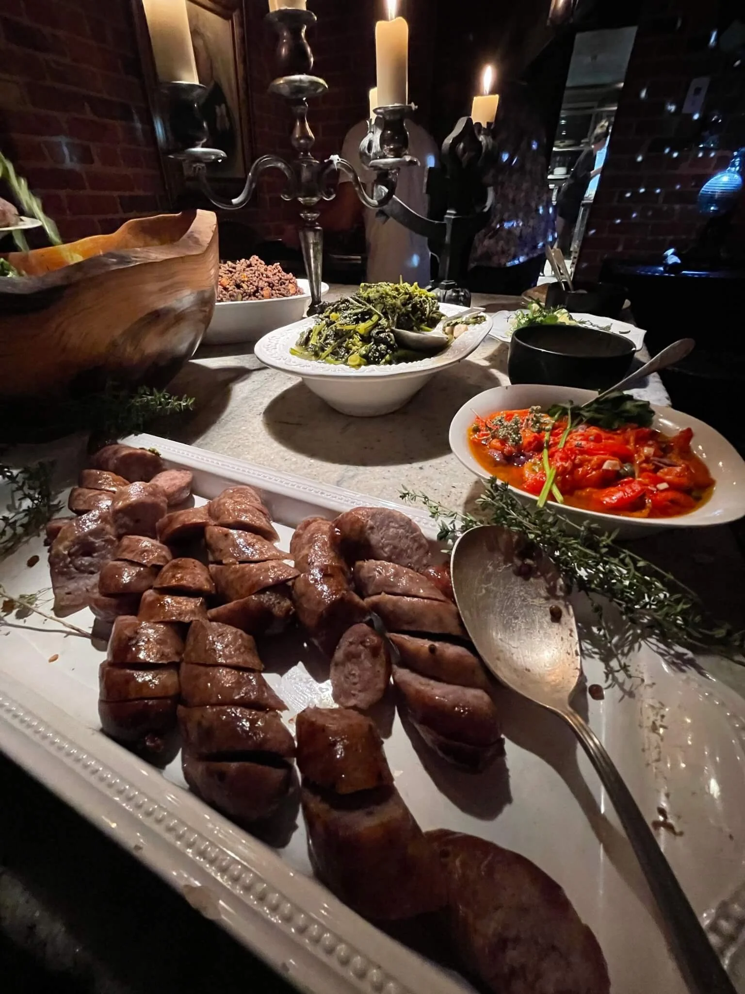 A table with various dishes including sliced sausage, a bowl of greens, a tomato-based stew or sauce, and a wooden bowl, with candlesticks and a brick wall in a dimly lit setting.