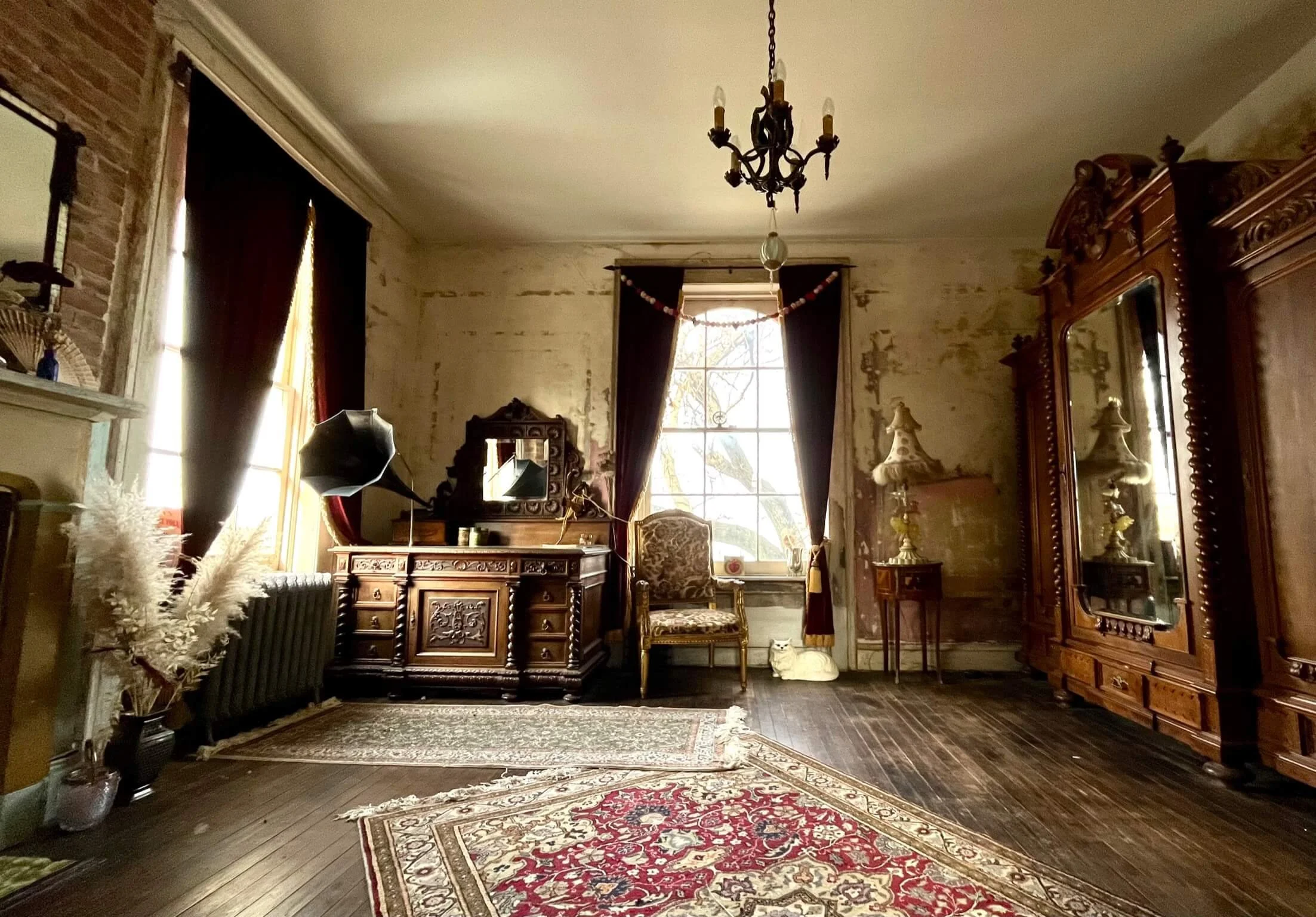 A vintage-style living room with antique wooden furniture, a chandelier, and a large window with dark curtains. The room has a patterned rug on dark wooden floors, a mirror, a chair, and decorative lamps.