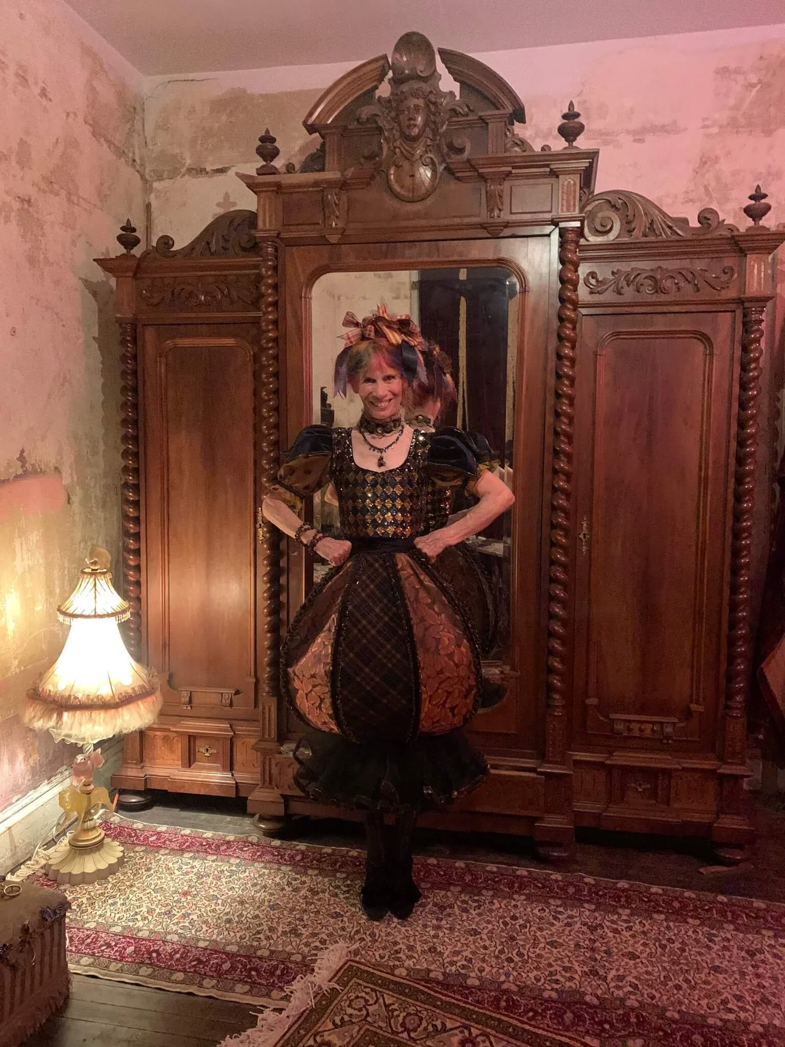 A woman dressed in a steampunk-inspired, elaborate, colorful Victorian costume with a large skirt, standing in front of an antique wooden wardrobe mirror in a vintage room with peeling walls and a patterned area rug.