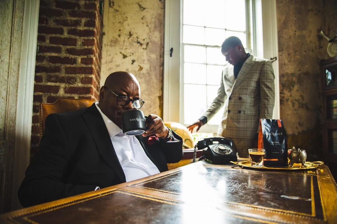 An older man with glasses in a suit sitting at a desk, drinking from a mug labeled 'KISS COFFEE'. A younger man in a checked blazer stands near a window with brick and textured walls behind. The desk features coffee bags, a phone, a glass of water, a