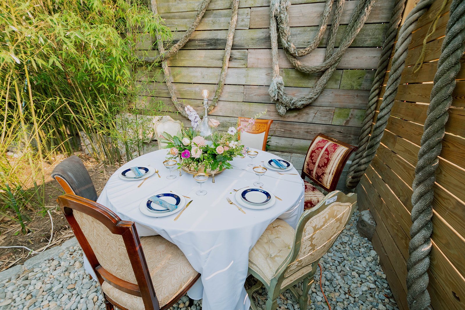 A round dining table set for six, with a white tablecloth and elegant tableware, in a rustic outdoor setting with wooden walls and greenery in the background.