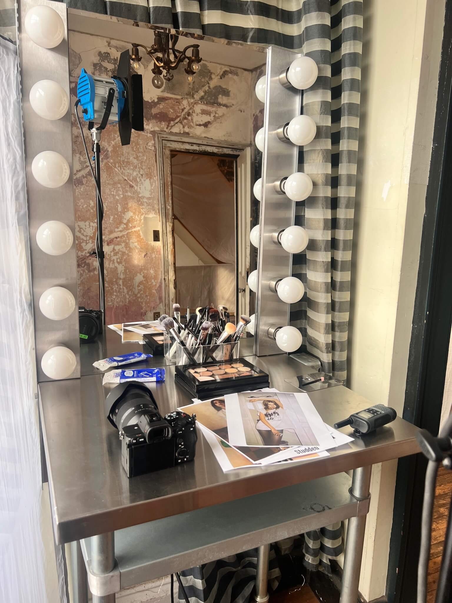 A makeup vanity table with a large mirror surrounded by light bulbs, makeup brushes, and cosmetic products. There is a camera and some printed photographs on the table. A camera and a remote are also visible on the table. The background shows a distr